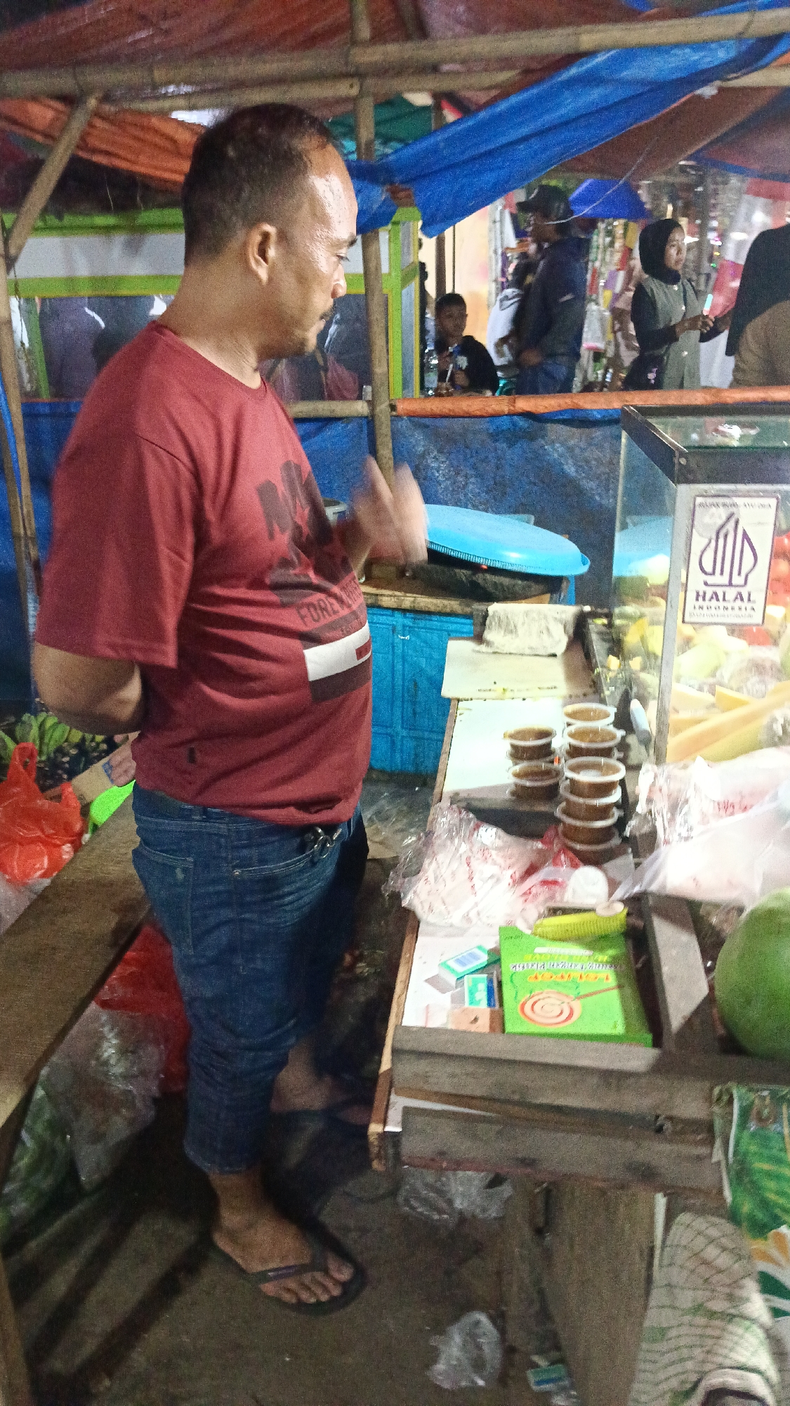 The image depicts an outdoor market scene in Tajurhalang, Indonesia. A man wearing a red t-shirt and blue jeans stands to the left, facing away from the camera. He appears to be gesturing with his right hand. Behind him, there are various stalls covered with tarps. In the foreground, to the right, a glass display case contains stacked servings of food, possibly sauces or condiments, and a green package with circular designs. A large green fruit, possibly a coconut, is visible on the right edge of the frame. The ground is dirt and debris. Several other individuals are visible in the background, blurred and out of focus, suggesting a busy market environment.