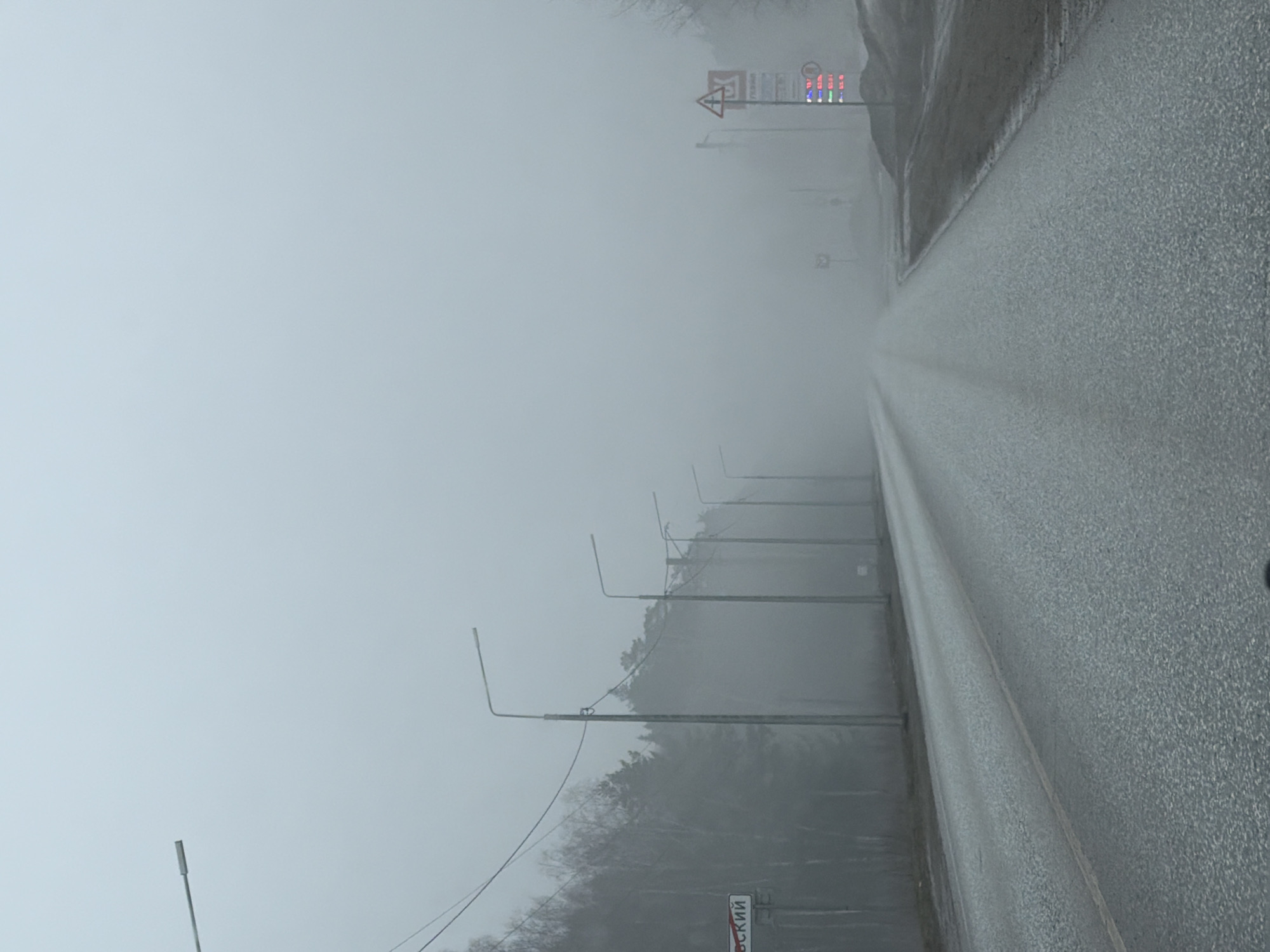 The image shows a straight, two-lane road disappearing into heavy fog. The road surface appears wet. On the right side of the road is a concrete barrier or wall. Streetlights line the left side of the road, spaced regularly, diminishing into the fog. In the far distance, a blurred sign is visible, displaying fuel prices and a hazard warning symbol. A road sign with Cyrillic letters is on the left side of the road, closer to the viewer. Given the Cyrillic letters and other details, it's in the city of Chaikovsky, Russia.