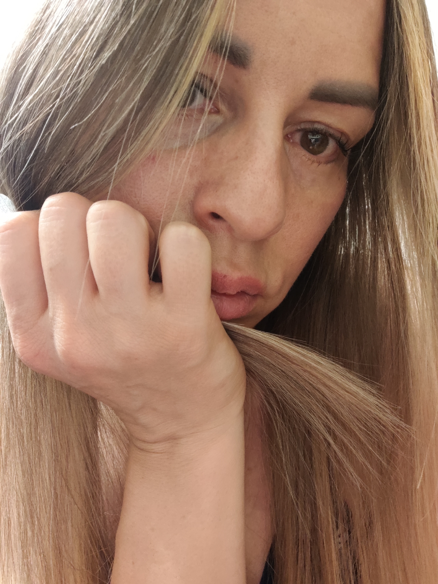 A woman, with long blonde hair, is shown in a close-up, chest-up, studio portrait. She has her right hand up to her face, with her fingers resting on her cheek. Her left hand is partially visible, holding some of her hair. The woman has brown eyes and is looking down to her right. The background is out of focus and appears to be a plain, light-colored wall. The image was taken indoors. The city of Almaty, Kazakhstan is not visible in this image.