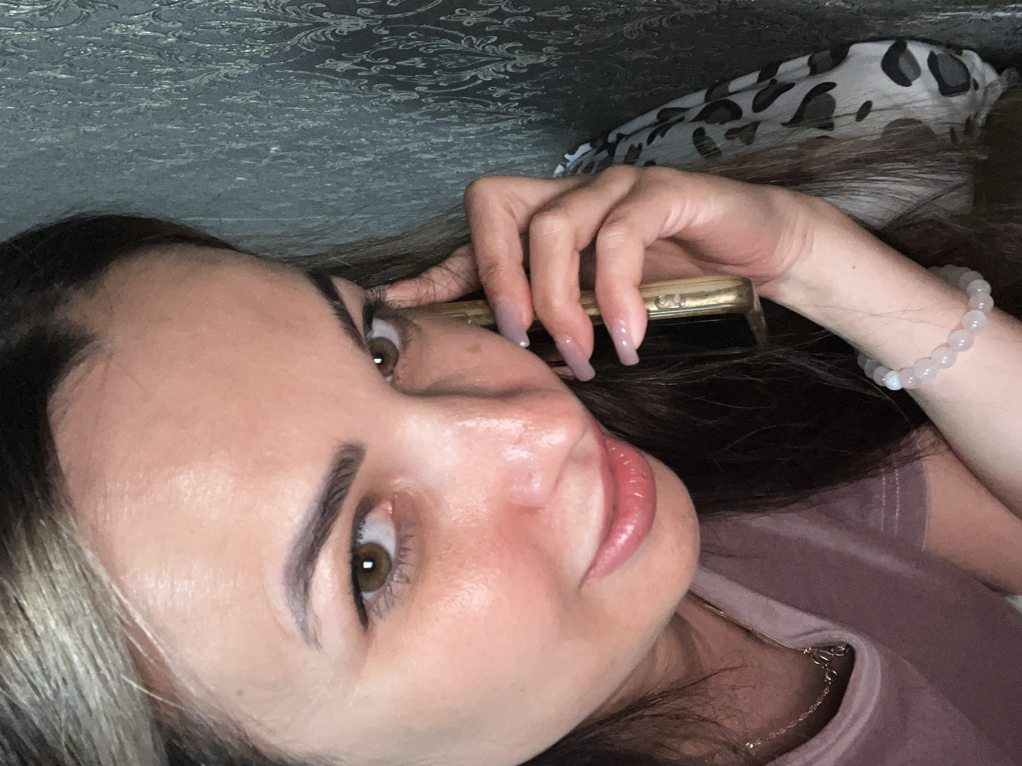 A woman with long, dark brown hair and light brown eyes is holding a gold-colored phone to her ear with her right hand. She is wearing a pale pink t-shirt and a delicate gold necklace with a small pendant. Her nails are long and light pink. She has a light dusting of makeup on her face, including mascara and lip gloss. The background appears to be a textured wall with a damask pattern. The lighting suggests an indoor setting, possibly a home.