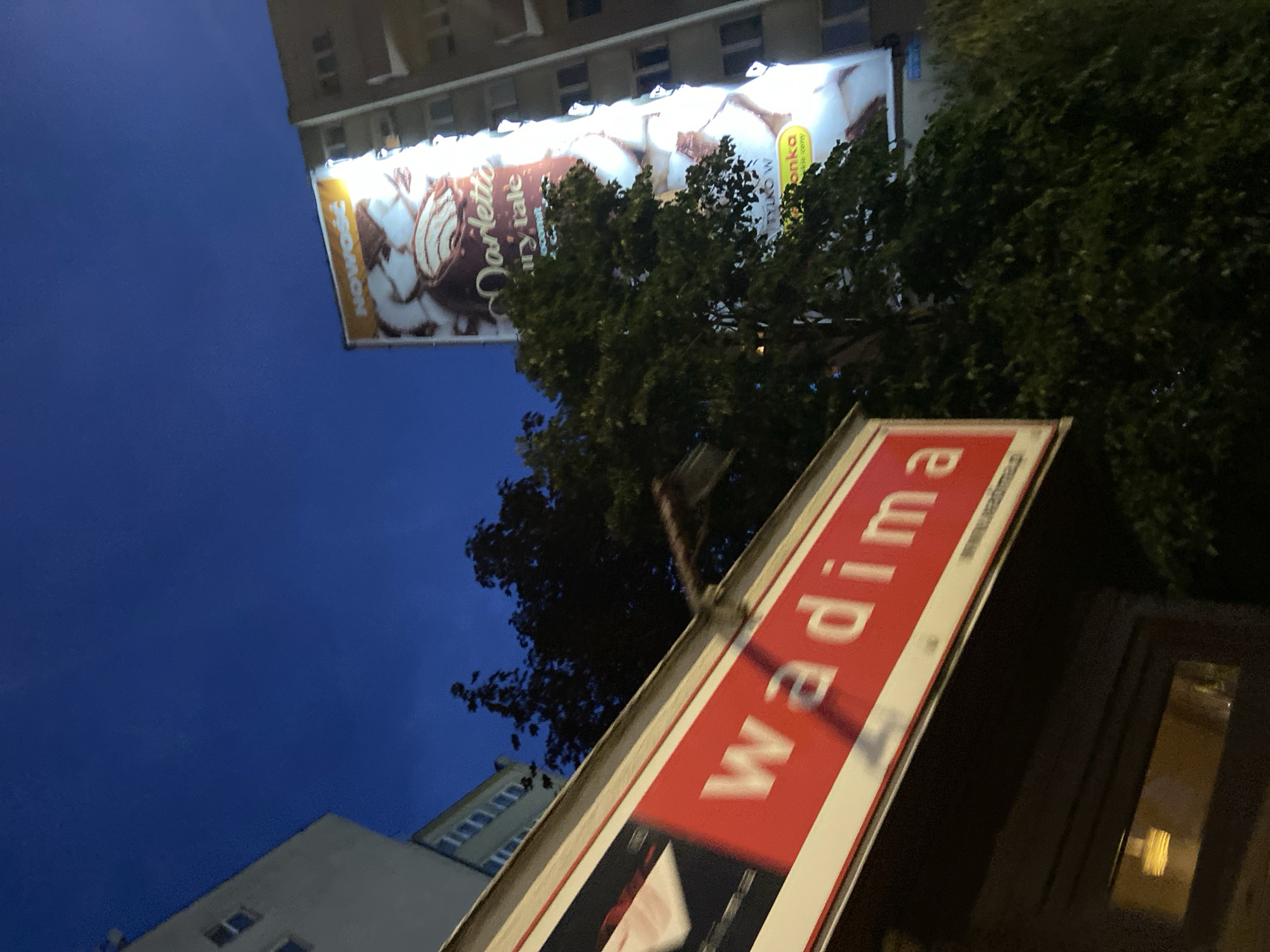 The image shows a street scene at dusk or night, with a deep blue sky visible. In the foreground, a red and white sign with blurred white text, likely a business name, is angled towards the viewer. Part of the text reads "wadima". Behind this sign, dark green trees obscure the view of some buildings. To the right, a tall, brightly lit billboard is mounted on the side of a building. The billboard displays an advertisement featuring a chocolate product and text in Polish, including "NOWOŚĆ" (New) and "Marletto". The building to the right of the billboard has multiple windows, some of which are lit. Another building, lighter in color, is visible on the left side of the image. The overall impression is an urban environment with commercial advertising illuminated against the night sky. This scene is located in Warsaw, Poland, on a street with multiple commercial establishments and residential buildings.