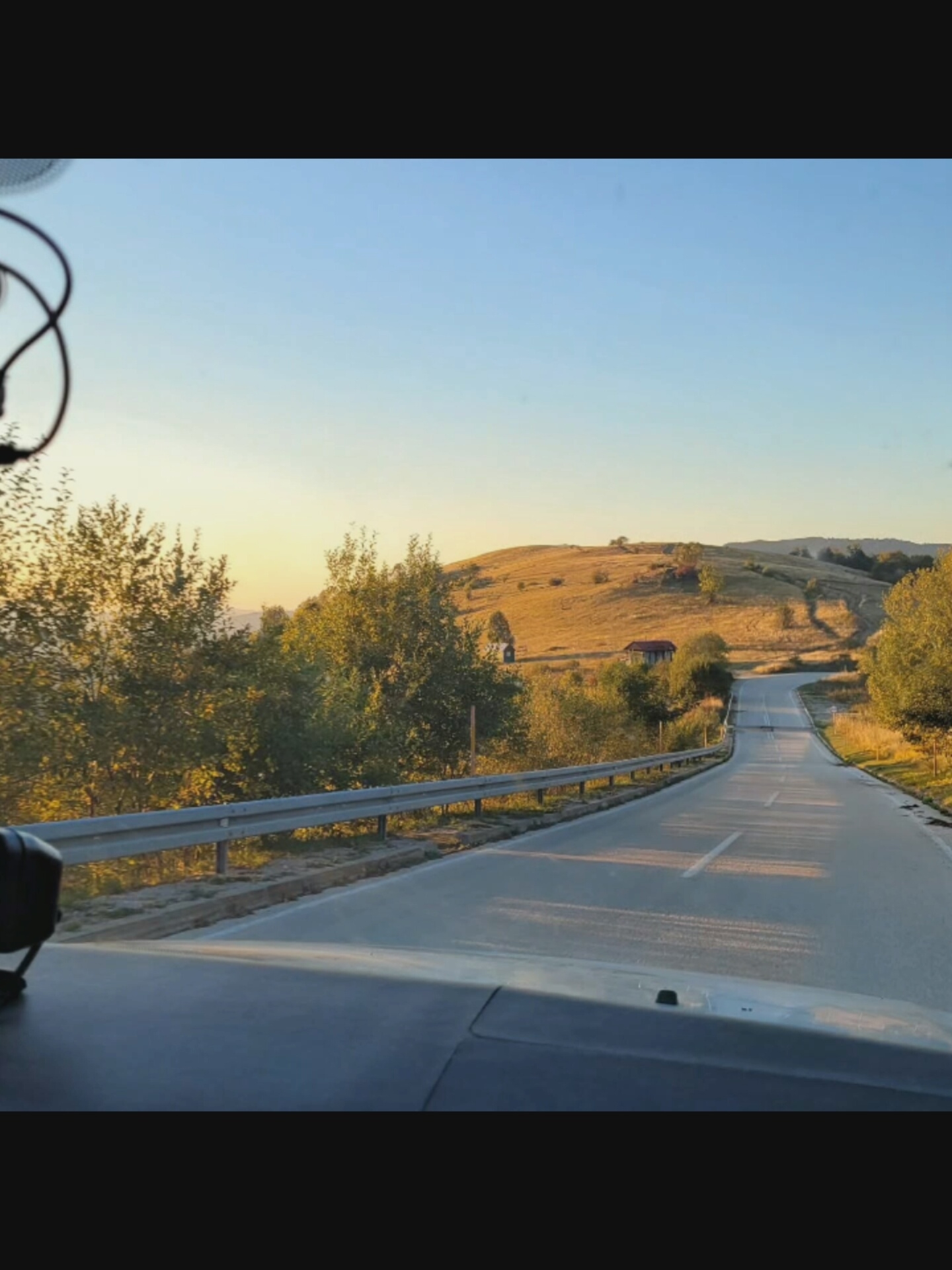 The image captures a view from inside a vehicle driving on a paved road that curves through a hilly, rural landscape. The time of day appears to be late afternoon or early evening, with the sun casting long shadows and illuminating the golden-brown hills and trees. The road has white lane markings. On the left side of the road, there is a metal guardrail. A few scattered trees and some small buildings, possibly houses or sheds, are visible on the hillsides. The sky is clear and blue, transitioning to a lighter hue near the horizon. There are no people visible in the image, and the focus is on the natural scenery and the road. The image does not depict any elements that would suggest it is located in the city of Bucharest, Romania; it appears to be a more rural setting.