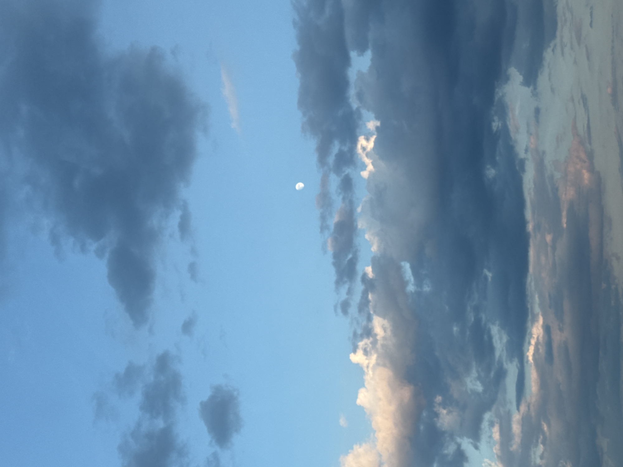 The image is a vertical, full-frame shot of a cloudy sky. The sky is a pale blue, with a few small, scattered dark clouds and larger, more dense dark clouds towards the bottom of the frame. There are also some lighter, wispy clouds, particularly in the lower half of the image, some of which appear to be illuminated with pinkish hues. A small, bright, partially illuminated moon is visible in the upper-middle section of the sky, off-center to the left. The scene depicts a natural outdoor environment. There are no people, man-made structures, or specific landmarks visible that would indicate its location within the city of Войводино, Bulgaria.