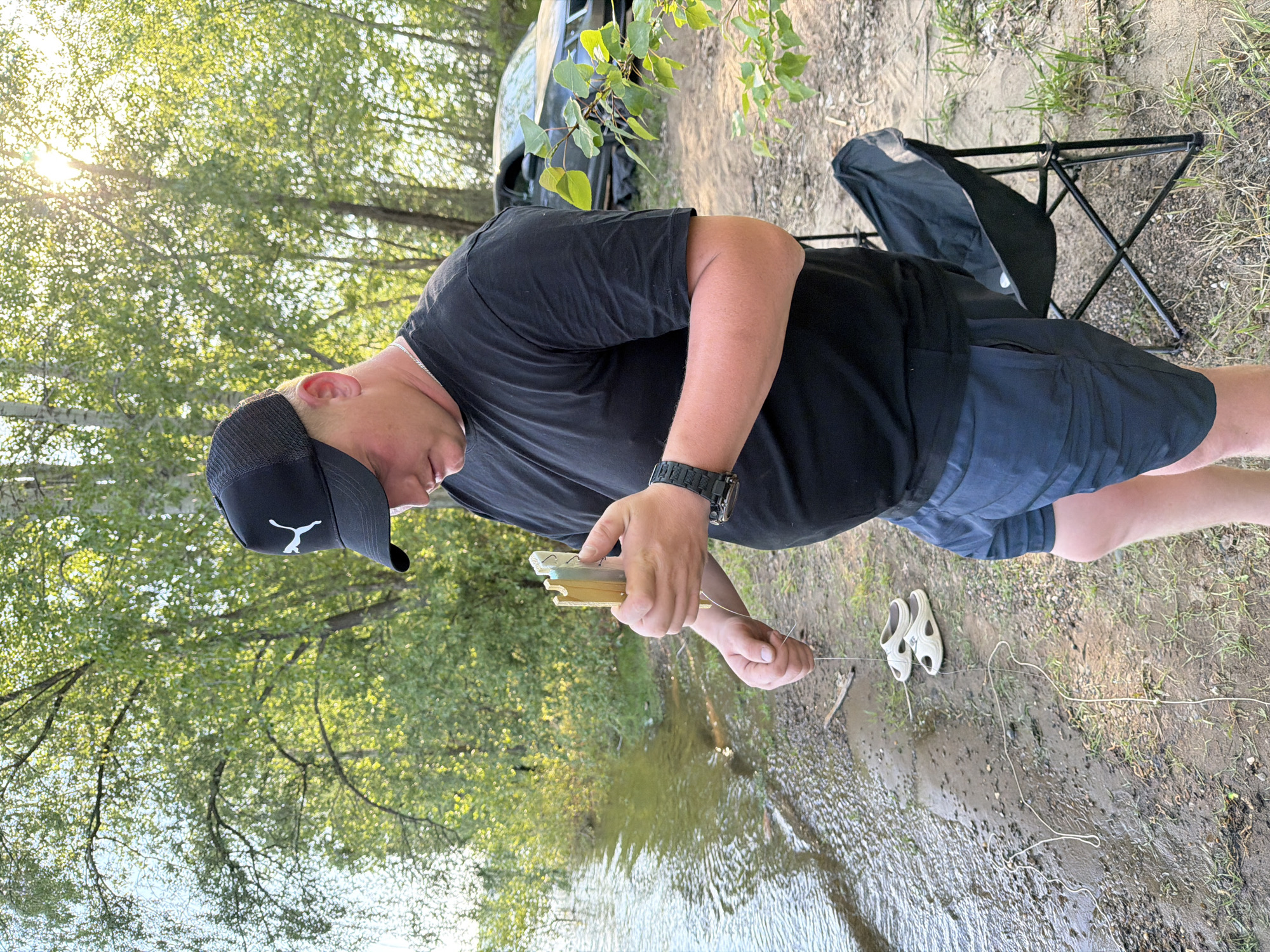 A man wearing a black t-shirt, dark shorts, and a black baseball cap with a white logo is standing on the edge of a body of water, possibly a river or lake. He is holding a small rectangular object with several fishing hooks attached to it. The man is looking down at the fishing line that extends from the object into the water. Behind him, there is a folding camping chair, and further in the background, a car is partially visible. The environment consists of trees and vegetation, suggesting a natural setting. There is no explicit information suggesting the location being Noviy, Russia.