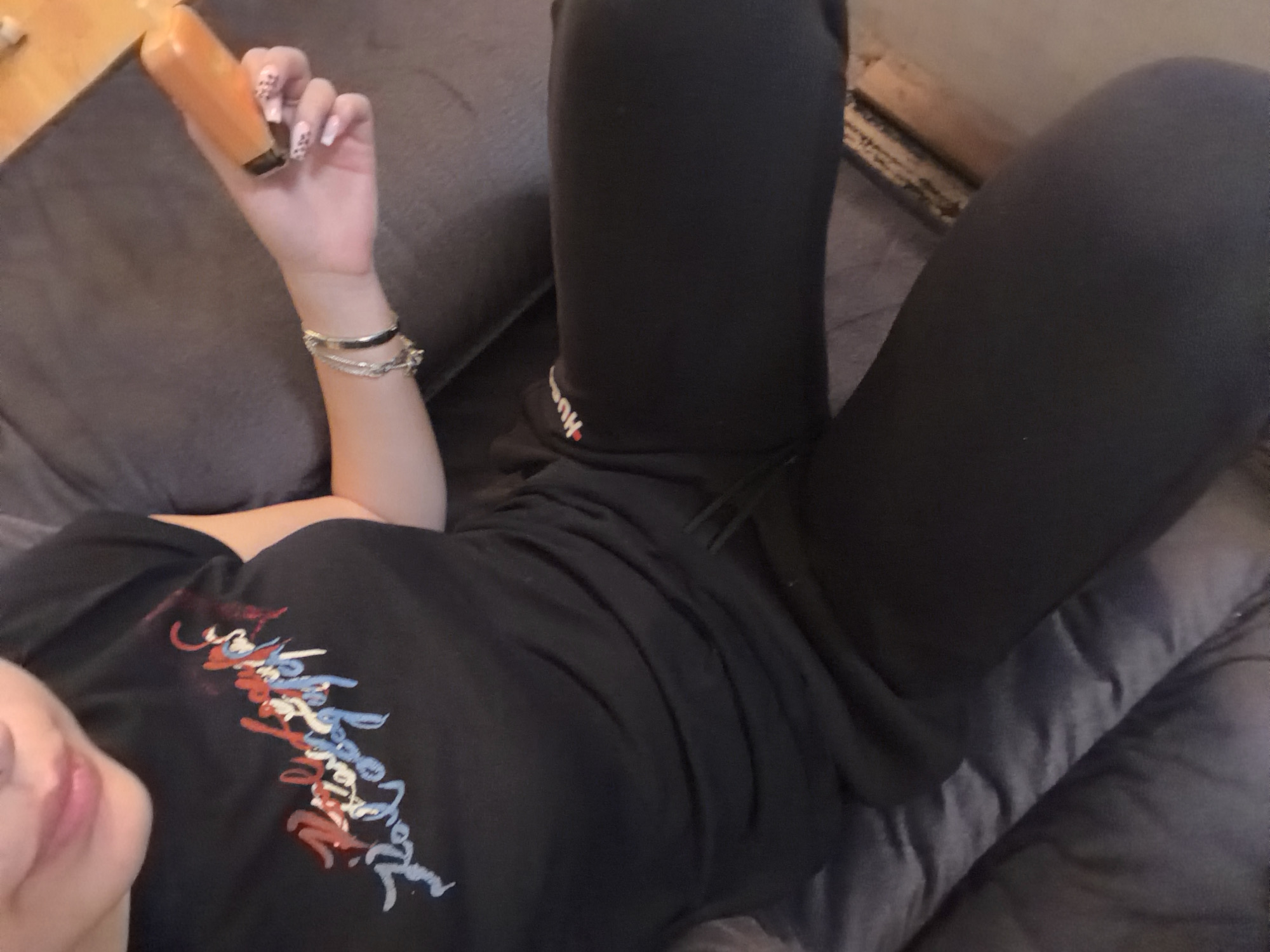The image shows a person lying on a gray couch. They are wearing black sweatpants and a black t-shirt with a colorful design on the front. The person is holding a small, rectangular object in their left hand and has bracelets on their wrist. The location is not evident from the image.