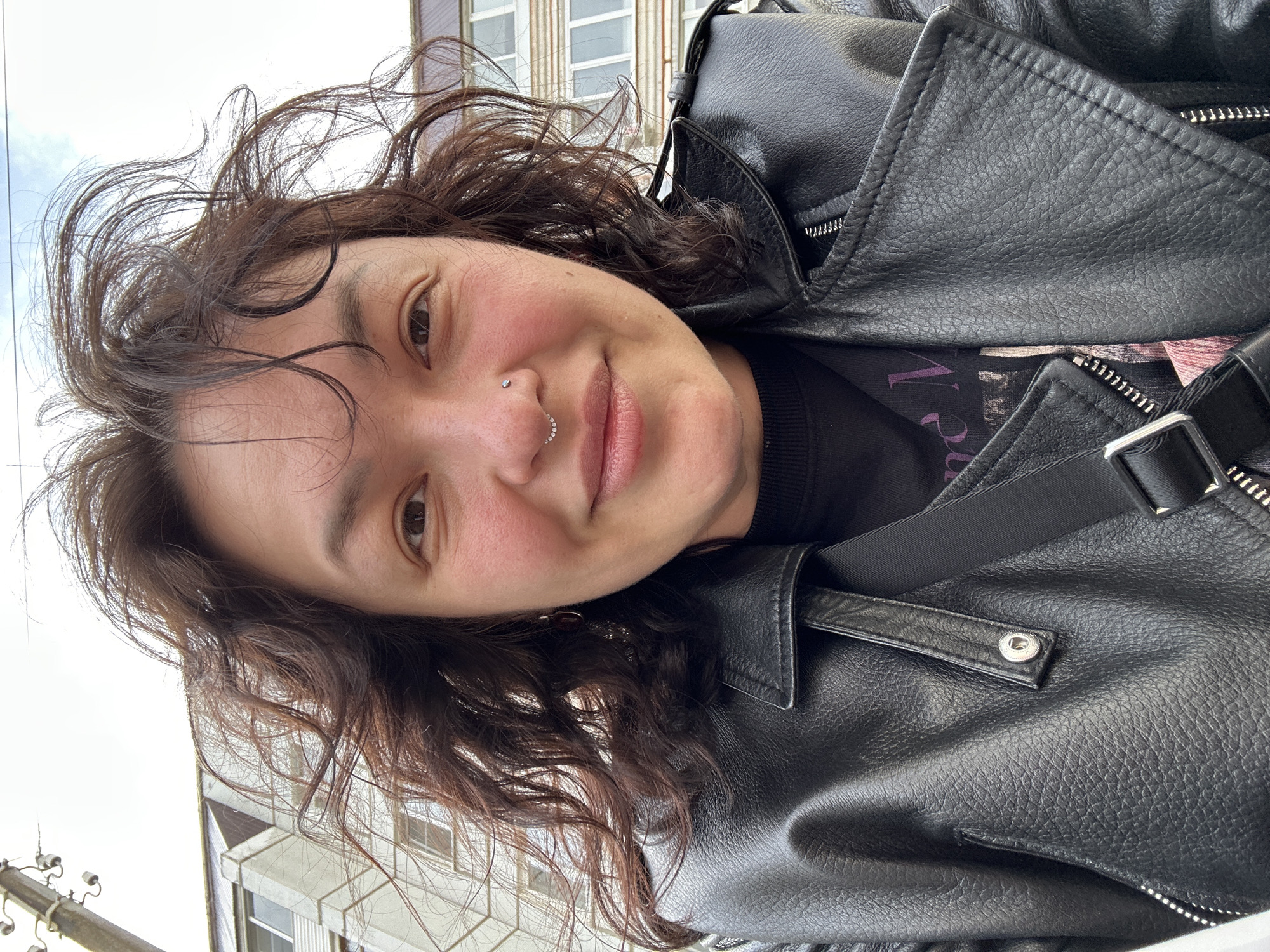The image is a close-up, tilted selfie of an individual, likely taken outdoors in Chaikovsky, Russia, during daytime under an overcast sky.
The main subject is an adult with medium-length, wavy dark brown hair, smiling warmly with closed lips and rosy cheeks. They are looking directly at the camera. The person is wearing a black leather-like jacket with visible zippers, snap buttons, and collar details. A dark, possibly black, long-sleeved top is worn underneath, with a black strap, likely from a bag, visible across the shoulder.
Notable details include a small silver stud piercing on the left nostril and a segmented ring, potentially adorned with small stones, on the lower lip.
The background shows elements of an urban or town environment, with parts of light-colored buildings featuring windows and one building having a brown trim near its roofline. A utility pole with wires or insulators is partially visible on the far left, further indicating an outdoor setting.
The overall emotion conveyed is one of happiness and approachability. Indistinct purple text, possibly starting with "V" (e.g., "Vie" or "Violet"), is faintly discernible on the dark fabric underneath the jacket collar.