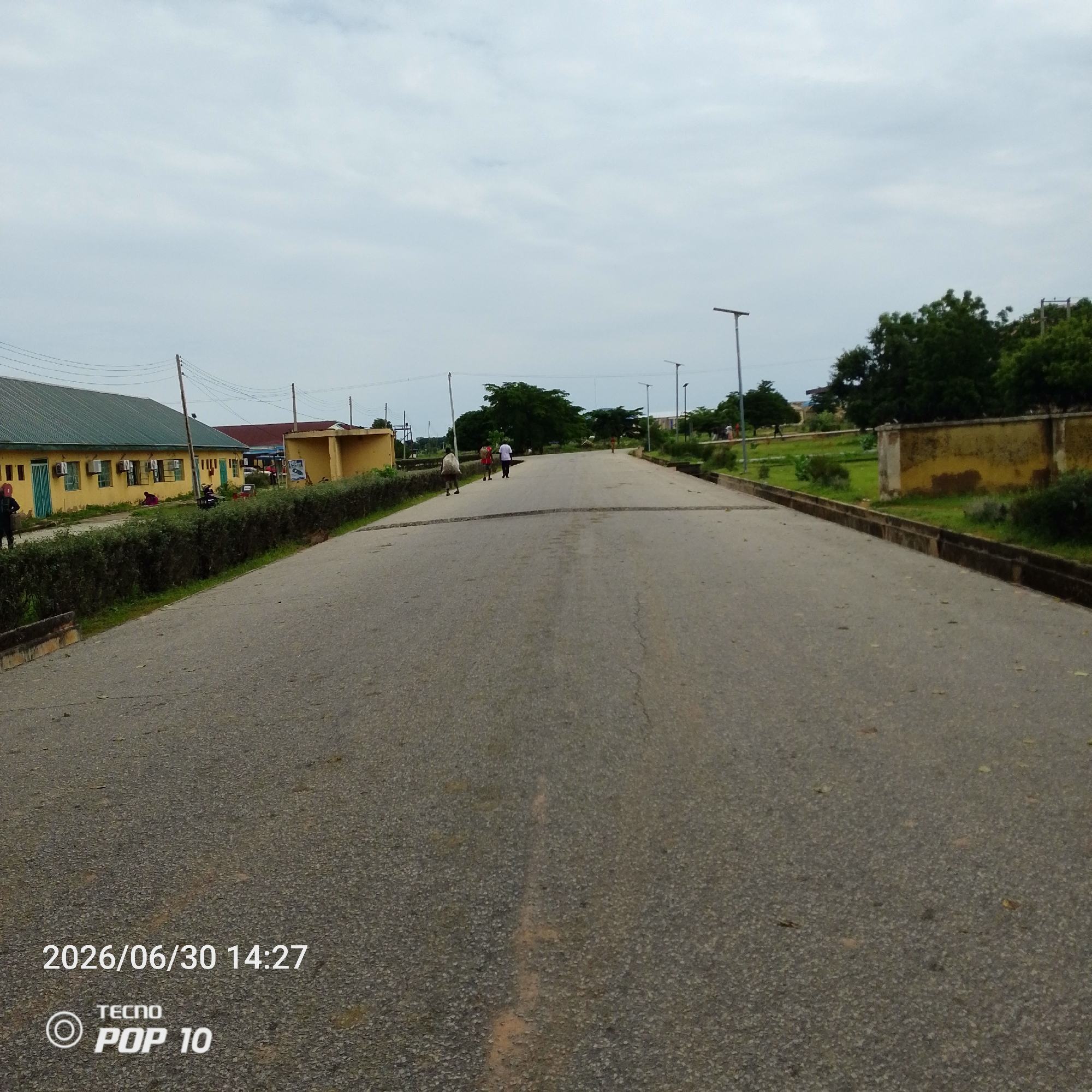 The image depicts a wide, paved road curving slightly into the distance under an overcast sky. On the left side of the road, a long, yellow building with a green roof is visible, featuring several windows and air conditioning units. A neatly trimmed green hedge runs along the edge of the building, separating it from the road. Further down the road, scattered people can be seen walking in both directions. Some appear to be carrying bags or bundles.
To the right of the road, there's a grassy area with trees and a low, weathered yellow wall. Several tall lampposts line the side of the road. The scene suggests a public space, possibly a campus or a community area, given the presence of buildings, manicured hedges, and the road infrastructure.
The lighting is diffused, indicating a cloudy day, and the time of day is noted as 14:27 on June 30, 2026, according to the visible text at the bottom left of the image. The text also indicates the device used to capture the image, "TECNO POP 10." No specific emotions are evident from the distant figures, and the overall atmosphere is that of a typical daytime scene in a location that appears to be Jalingo, Nigeria.