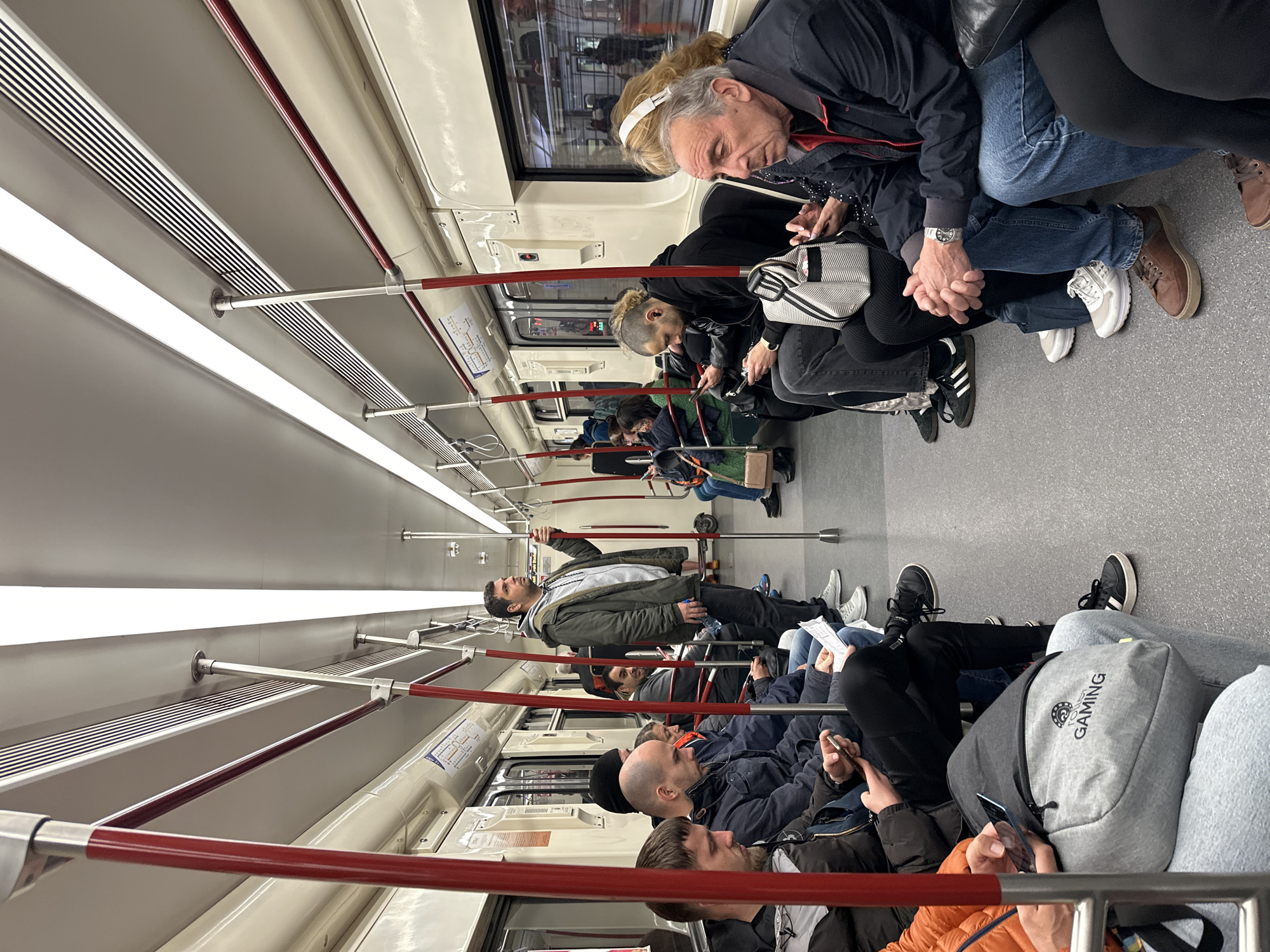 The image captures the interior of a metro train car in Sofia, Bulgaria, depicting numerous passengers during what appears to be a daily commute. The train carriage is brightly illuminated by long, overhead fluorescent lights, with a clean, modern design featuring light-colored walls and ceiling, and a gray floor.
Most of the visible passengers are adults, primarily seated along the left side of the carriage. Many individuals are engrossed in their smartphones, looking down at their screens. In the foreground, an older man with light hair and a white headset is seated, focused on his device. Next to him, a person with short, distinctive hair is also using a phone. Further down the carriage, other passengers are seated, some reading or simply resting. A notable detail is a pair of hands clasped together, suggesting a couple sharing the journey.
On the right side of the carriage, a man in a dark green jacket stands, holding onto a vertical handrail, with a water bottle in hand. Behind him, more seated passengers are visible through the train's sturdy red and metallic handrail system that runs horizontally and vertically throughout the car, providing support. The train's interior also features ventilation grilles and window panels.
Visible text includes "100% GAMING" and a circular logo on a gray backpack in the lower right portion of the image. The Adidas logo is visible on several pairs of sneakers worn by passengers. Small informational signs or route maps are affixed to the walls but are unreadable. The overall scene portrays a typical moment of urban transit, with commuters quietly occupying their journey.