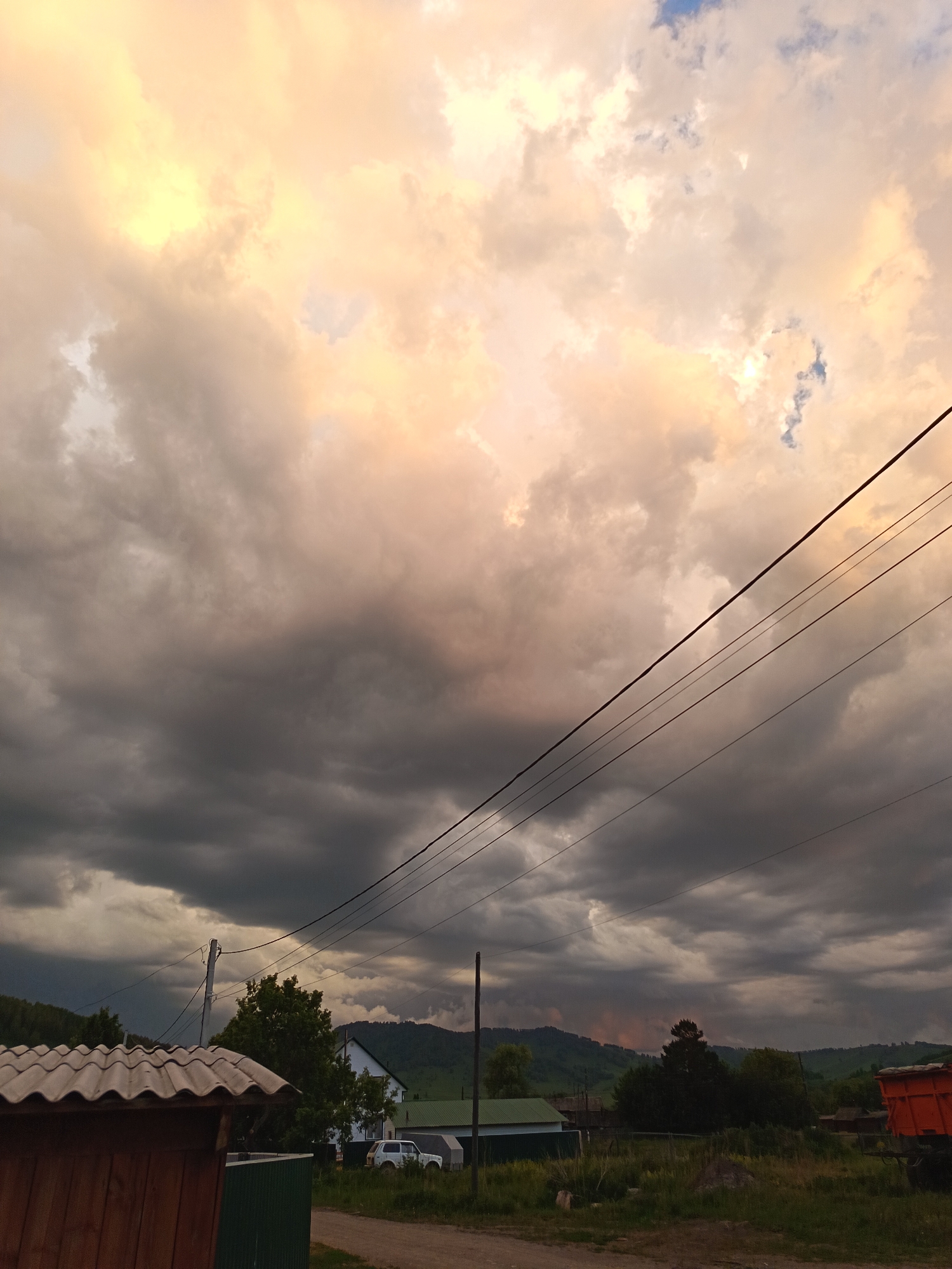 A photograph captures a village scene in Soloneshnoye, Russia, under a dramatic sky. The sky is filled with a mix of dark, heavy clouds and patches of bright, warm light, suggesting either the end of a storm or an approaching one. Power lines stretch diagonally across the frame, bisecting the sky.
In the foreground, on the left, is the corner of a wooden structure with a corrugated roof. To its right, a green metal fence is visible. Beyond these structures, a dirt road leads into the scene.
The middle ground features several buildings. A white house with a dark roof stands prominently, partially obscured by trees. To its left, a smaller building with a green roof is visible. A white SUV is parked near the white house. Further into the background, rolling hills covered in trees form the horizon line. The sky above these hills is also filled with the same turbulent cloud formations.
On the far right of the image, the back of an orange truck with a load is visible, suggesting it is parked or moving slowly. The overall impression is one of a rural landscape at dusk or before a storm, with natural elements and human structures coexisting under a dynamic sky.