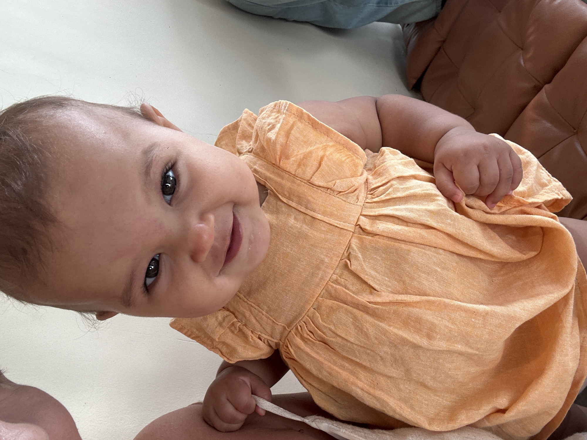 A baby wearing an orange dress looks up towards the camera. The baby is sitting on someone's lap, holding a white cloth. The background consists of a light-colored surface and a brown leather couch. This image was likely taken indoors. There is no information available to indicate the image's location in North Kuta, Indonesia, or if it was even taken there.