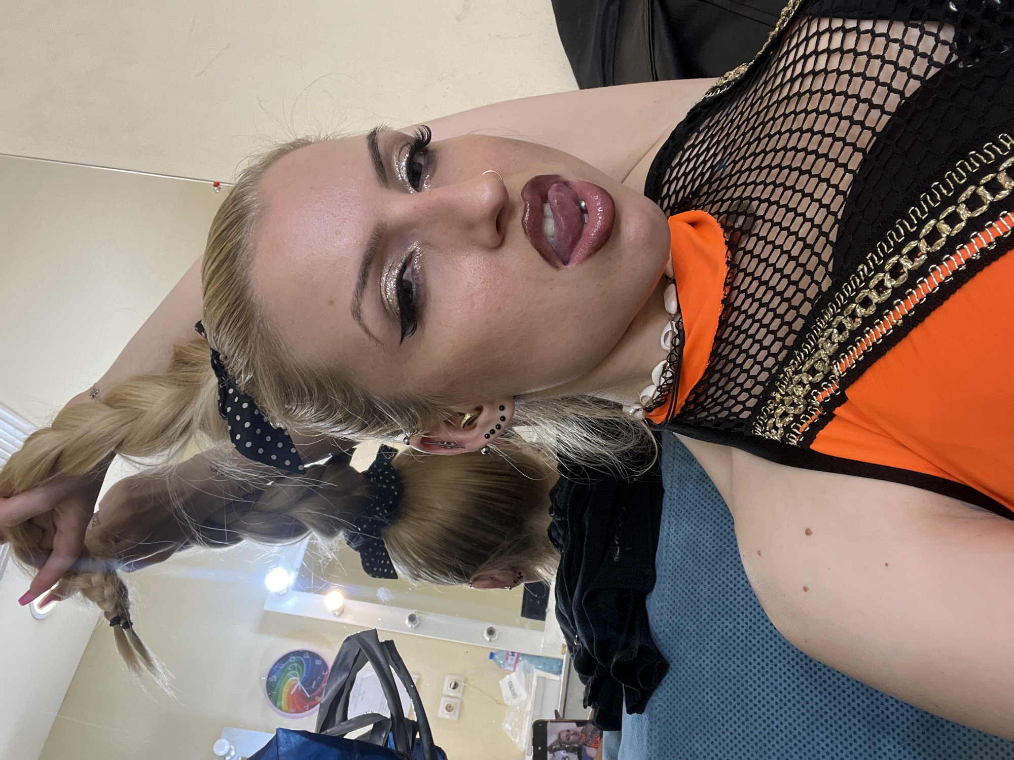 A woman with blonde hair is in a room with a vanity mirror and lights in the background. She is wearing a black and orange outfit with a fishnet top, a nose ring, and glittery eyeshadow. Her hair is styled in a braid with a black polka dot scrunchie. She is looking at the camera with her tongue out. There is a phone on a blue surface with a blurry image of the woman on the screen. There are bags on the floor. It's impossible to confirm the location in Plovdiv, Bulgaria, based on the visible content of the image.