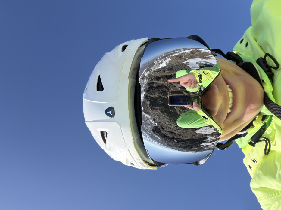 The image displays a close-up, low-angle perspective of an individual dressed in winter sports attire against a clear blue sky, located in Zelenaya Polyana, Russia. The subject wears a white ski helmet, bearing an "ATOMIC" brand label on the left side and a triangular logo centrally. Large, reflective, mirrored ski goggles obscure the upper face. The lower portion of the subject's face, from the nose to the chin, is visible beneath the goggles, showing a slight smile with upper teeth exposed. A neon green ski jacket with black detailing around the collar and chest is visible. The prominent feature of the image is the detailed reflection within the goggles, which reveals a rugged, mountainous landscape composed of dark, rocky terrain partially covered with snow. Within this reflection, the subject's left hand is visible holding a dark smartphone, indicating a selfie being taken, while the right hand displays a "thumbs up" gesture. The reflected attire of the individual also appears neon green. The environment is characterized by a bright, cloudless blue sky, suggesting clear weather conditions.