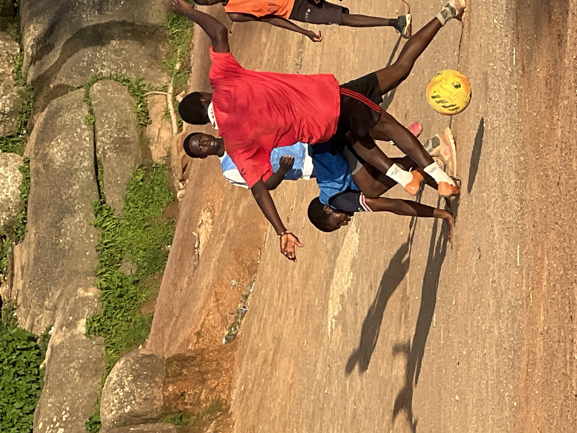 In Jos, Nigeria, several young men are playing soccer on a dirt field in front of large rock formations covered in green vegetation. The lighting suggests it is late afternoon, casting long shadows across the ground. One player in a red shirt and black shorts is kicking a yellow soccer ball, while another player in a blue shirt is attempting to block him. Two other players are visible in the background, one in a blue and white shirt and another in an orange shirt. The ground is a reddish-brown dirt, and scattered rocks and pebbles are visible. The overall scene depicts a casual outdoor soccer game in a natural, rocky environment.