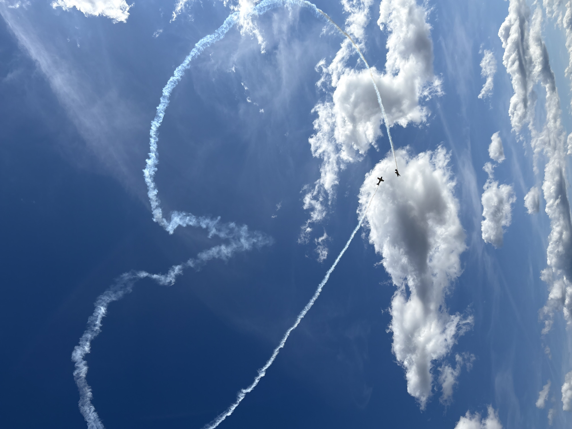 In the skies above Kynšperk nad Ohří, Czechia, two planes are performing an aerial display, leaving behind a heart-shaped contrail. The planes are small, dark silhouettes against the bright blue sky, with wisps of white smoke trailing from their wings, forming the distinctive heart shape. Fluffy white clouds are scattered across the sky, adding texture and depth to the scene. One plane is positioned slightly ahead of the other, both flying in formation. The contrail itself is a prominent feature, curving gracefully across the frame and indicating the path of the aircraft. The overall impression is one of a vibrant sky performance.