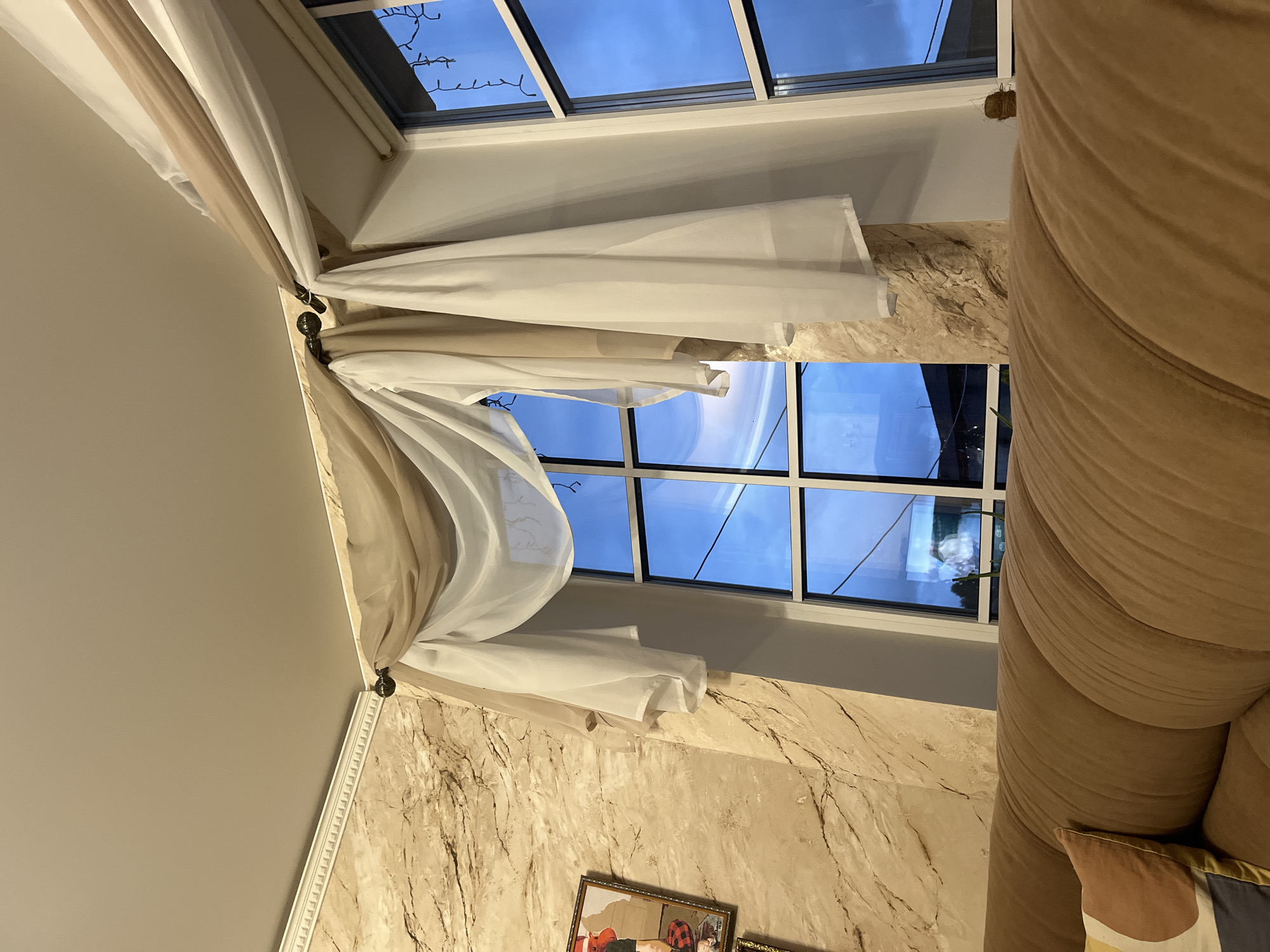 The image depicts an interior scene with a focus on a window dressed with sheer, layered curtains. The curtains, in shades of beige and white, are draped over a dark curtain rod. Through the window, a dusky blue sky is visible, reflecting the evening light. The window is divided into multiple panes by white mullions. To the left of the window, a wall with a textured, marble-like finish in shades of beige and brown is visible. A framed picture hangs on this wall, partially visible in the left portion of the image. In the foreground, the top of a brown sofa is visible, with a cushion featuring a geometric pattern. String lights are draped along the top of the window on the right side. The scene appears to be in a residential setting, likely a living room. There are no individuals present in the image, and no actions are being performed. The environment is indoors, with the exterior visible through the window. The location is stated as Vladimirovka, Russia.