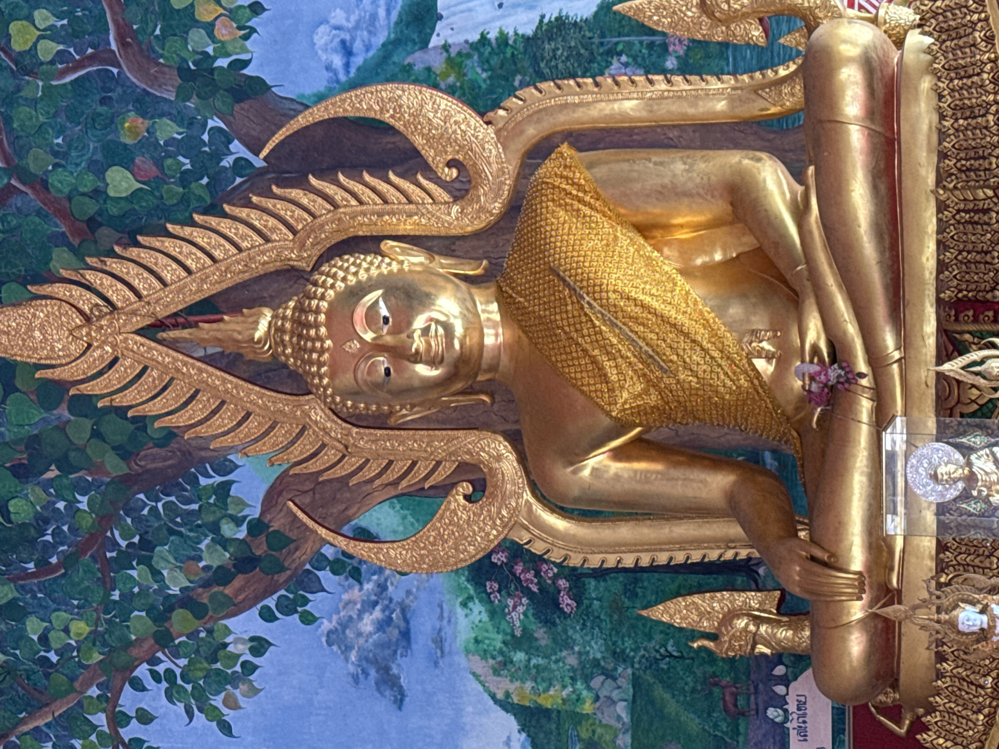 This is a close-up, eye-level shot of a golden Buddha statue seated in a meditative pose. The statue is adorned with a golden robe and is positioned in front of a mural depicting a lush landscape with a tree and mountains. The mural features a tree with green leaves, a body of water, and a waterfall in the background. There are also some smaller decorative elements at the base of the statue, including intricate golden carvings and what appear to be smaller Buddha images in glass cases. The overall impression is one of serenity and reverence. The statue is likely located in a temple or religious site in Ko Samui District, Thailand, given the style of the statue and the mural.