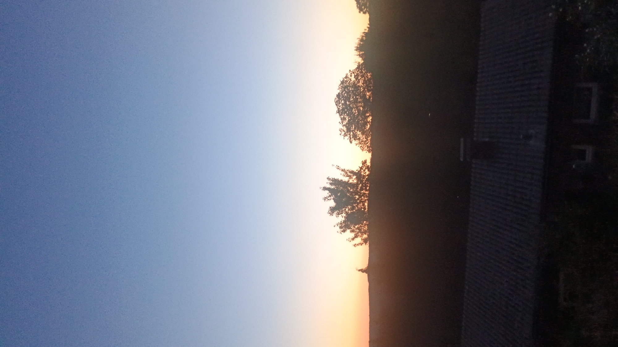 An outdoor, medium shot captures a sunset over a rural landscape, likely in Windeck, Germany. The sky transitions from a pale blue at the top to a soft orange and yellow hue near the horizon. Silhouetted against the fading light are trees of varying sizes and shapes, with a dark, undulating line of foliage separating the sky from the foreground. In the lower part of the image, the dark, sloped roof of a building with corrugated tiles is visible, featuring a chimney. The scene is dimly lit, suggesting either early morning or late evening.