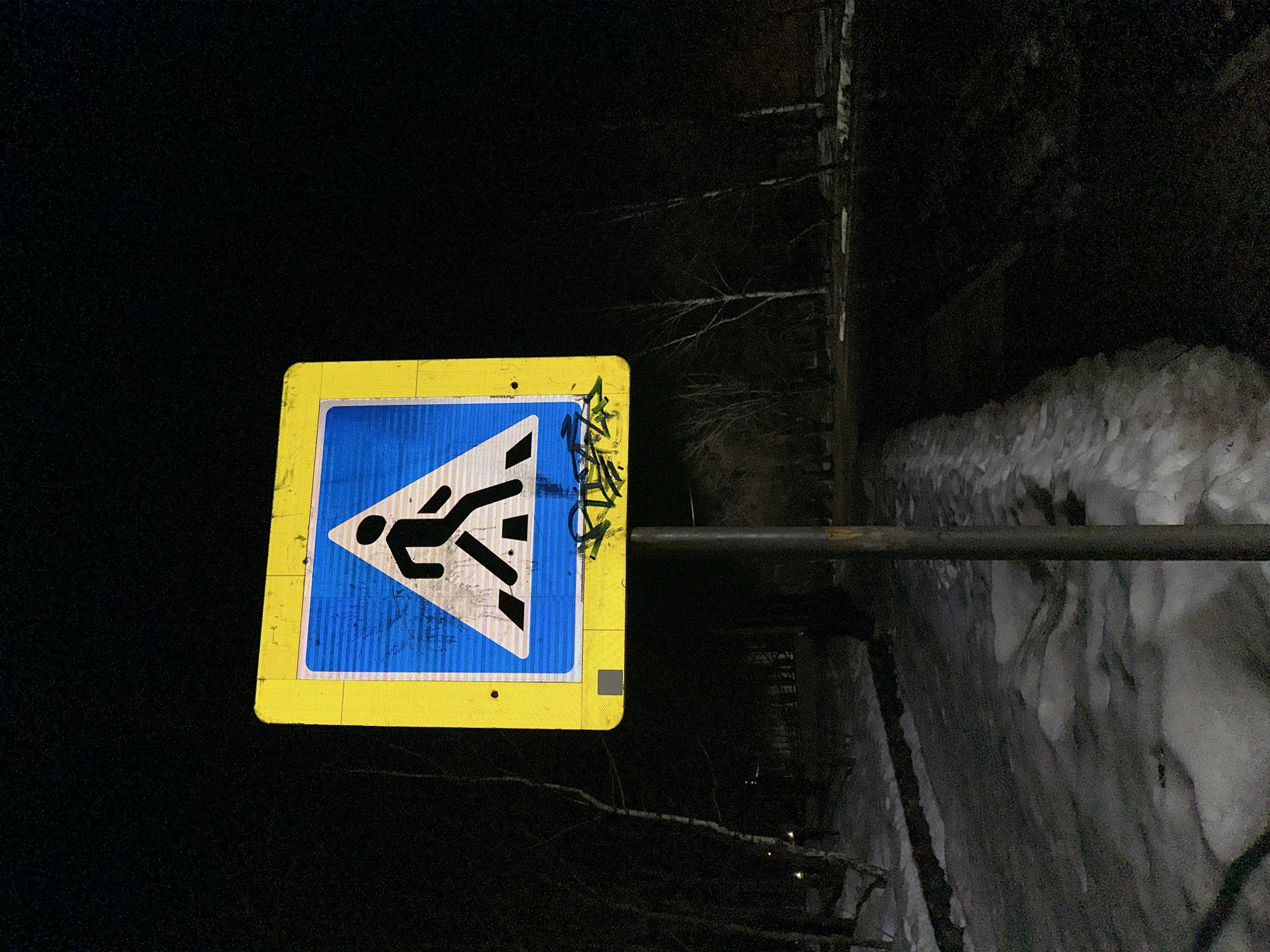 A pedestrian crossing sign is prominently displayed in the Sarapul, Russia scene. The sign has a yellow border and a blue interior featuring a black pedestrian silhouette within a white triangle. Graffiti partially obscures the upper-right corner of the sign. The sign is mounted on a metal pole. The surrounding environment is dark, with snow visible on the ground adjacent to what appears to be a road or path. Bare trees are visible in the background.