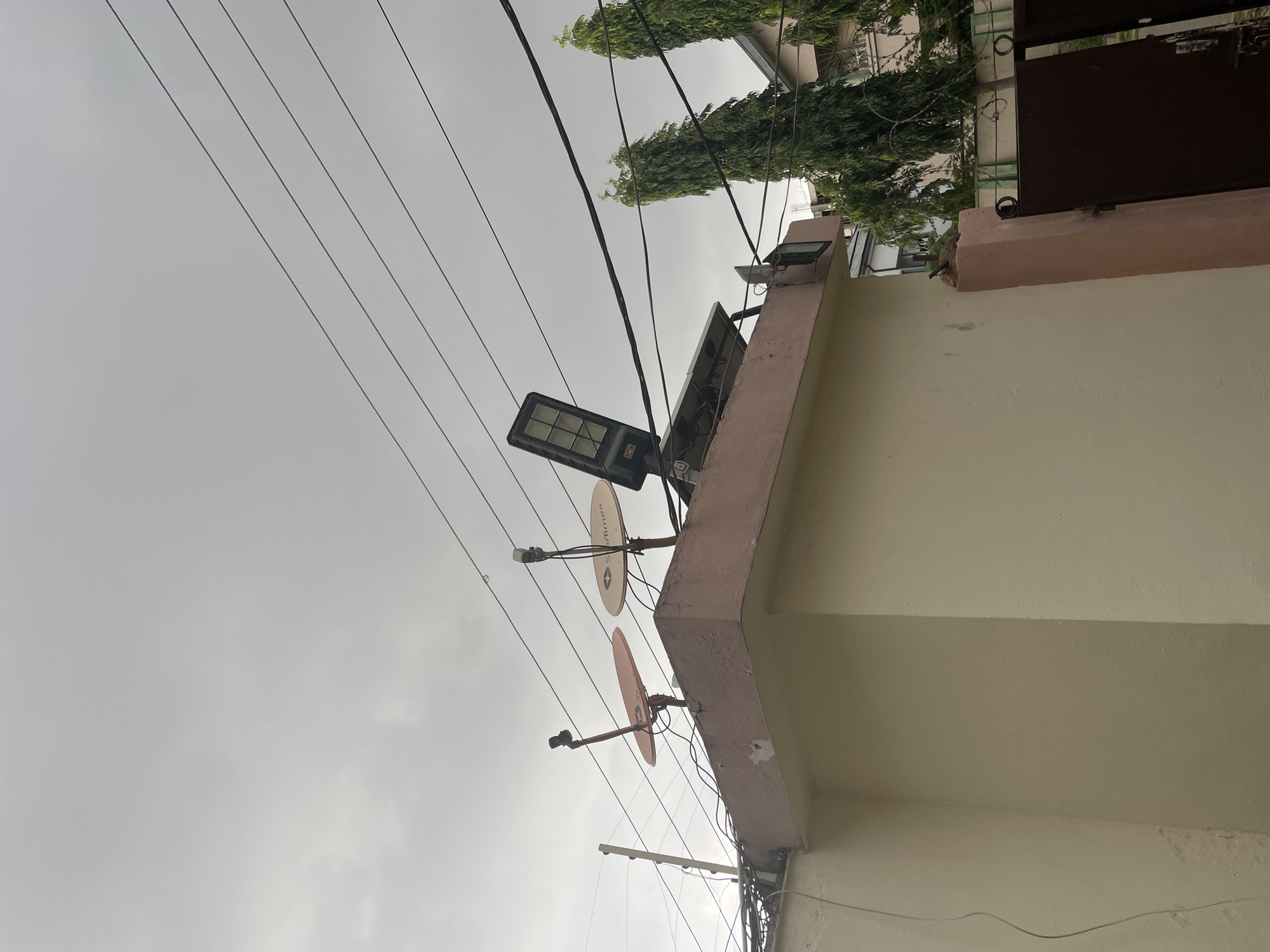 This image shows the top of a cream-colored building in Abuja, Nigeria, on an overcast day. Several satellite dishes, a solar panel, and a large LED light fixture are mounted on the roof. Power lines crisscross the sky above. To the right of the building, two tall, slender green trees are visible, with other buildings in the background. A dark metal gate is partially visible in the lower right corner.