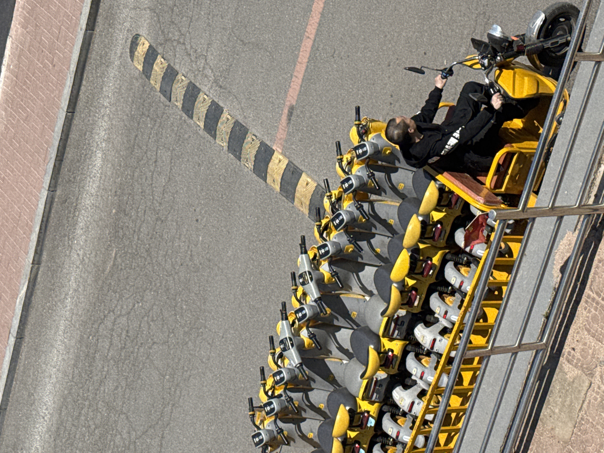 A high-angle view shows a collection of yellow and gray electric scooters parked in a neat row, likely for shared public use, adjacent to a street in the city of Zhongtun, China. A man dressed in black is sitting on the first scooter in the row. A speed bump, painted with black and yellow stripes, is visible on the road beside the scooter parking area. The environment appears to be a well-maintained urban area with visible paving stones and road markings.