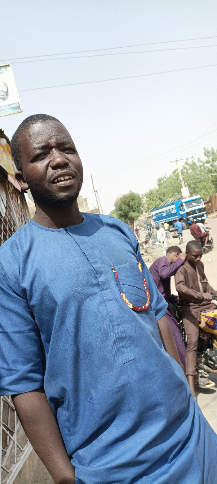 The image displays a street scene in Kaura Goje, Nigeria, under bright, clear daylight with a pale blue sky overhead, crossed by utility lines. The central figure is an adult male with dark skin, short dark hair, and a beard, positioned in the foreground, looking upwards and to the right with a slightly open mouth. He wears a solid blue, long-sleeved tunic, which features a left chest pocket adorned with a colorful fabric trim displaying red, yellow, blue, and black patterns. His right arm hangs beside him.
In the midground, to the right of the central figure, a young male in a brown long-sleeved garment sits on a yellow and black motorcycle. Another male, wearing a purple long-sleeved garment, is partially visible nearby, leaning forward. Additional indistinct figures and bicycles populate the background, indicating active street traffic. On the far left, a partial white sign with "BASHIR ALI AHMED" visible hangs from a structure. Further back, a large blue and white truck with a distinct cargo area is visible on the street. Utility poles and sparse green trees are present throughout the background, with the ground appearing to be a mix of dirt and pavement.