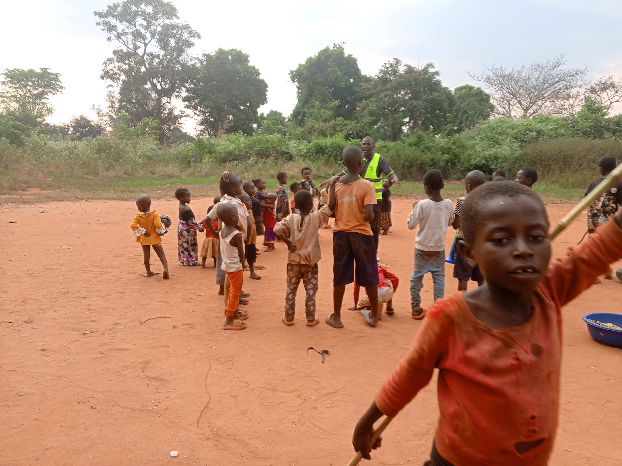 The image depicts a group of children and one adult in Ngalula, Democratic Republic of the Congo. The setting is an open, dirt-covered area with trees and vegetation in the background. The sky is overcast.
The adult wears a neon yellow vest and stands among the children. The children, varying in age and dress, are gathered in a group. In the foreground, a young boy is prominently featured, holding a stick and looking toward the viewer. A blue basin sits nearby. Other children are scattered throughout the scene, some interacting with each other. Clothing worn by the children appears worn and simple.