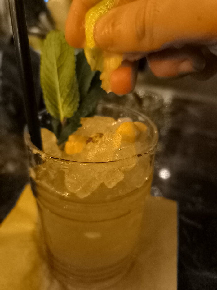 This close-up image shows a refreshing drink being prepared or enhanced in an indoor setting, likely a bar or restaurant, in Palaio Faliro, Greece. The central subject is a clear glass filled with a vibrant yellow-orange beverage, packed with crushed ice and small pieces of fruit, possibly orange or mango. A black straw is positioned on the left side of the glass, and a fresh sprig of green mint leaves serves as a garnish.
In the upper right portion of the frame, a hand with an orange hue is squeezing a halved citrus fruit, appearing to be a lemon or lime, directly over the drink. Droplets are visible falling from the fruit as it's being pressed, indicating the addition of fresh juice. The glass rests on a folded yellow or orange napkin or coaster, which blends with the color of the drink. The background is dark and appears to be a reflective surface, possibly a bar counter or table, with subtle highlights. The lighting is dim and atmospheric, suggesting an evening or nighttime setting. There is no visible text in the image.