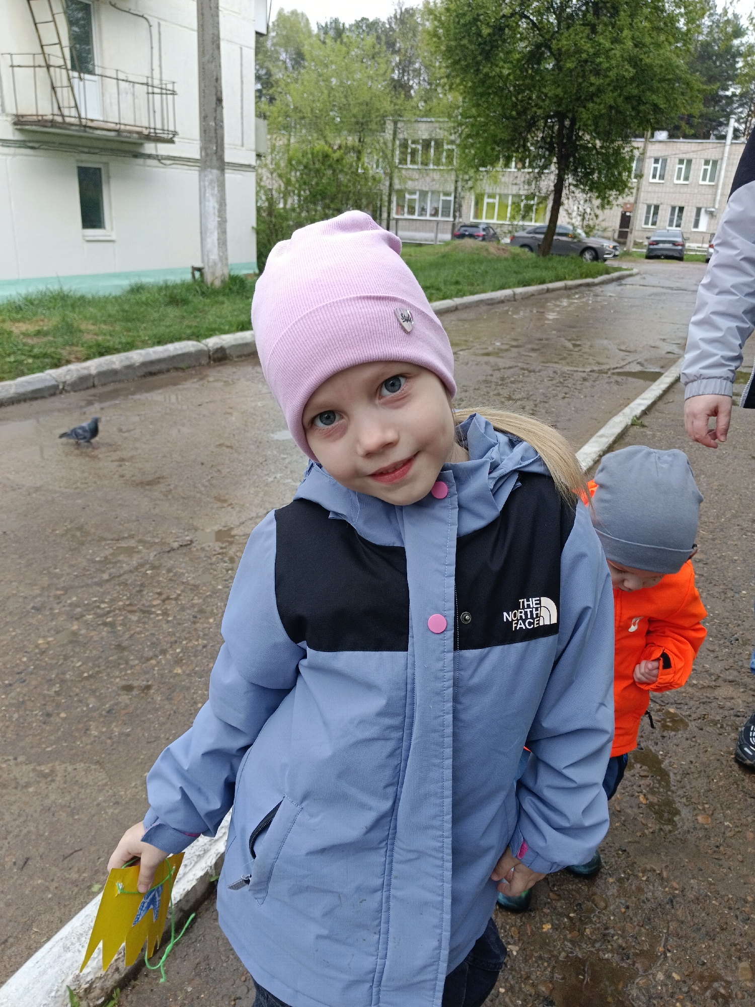 A girl in a pink hat and blue jacket is in a wet outdoor area, possibly a street, in Chaikovsky, Russia. She is holding a yellow object in her left hand. To her right, a younger child in a grey hat and orange jacket is visible. An adult is partially visible to the right of the younger child, wearing a grey jacket. A pigeon stands on the wet pavement to the left of the girl. In the background, there are apartment buildings and trees, suggesting an urban or suburban environment. The area appears to be wet, possibly after rainfall.