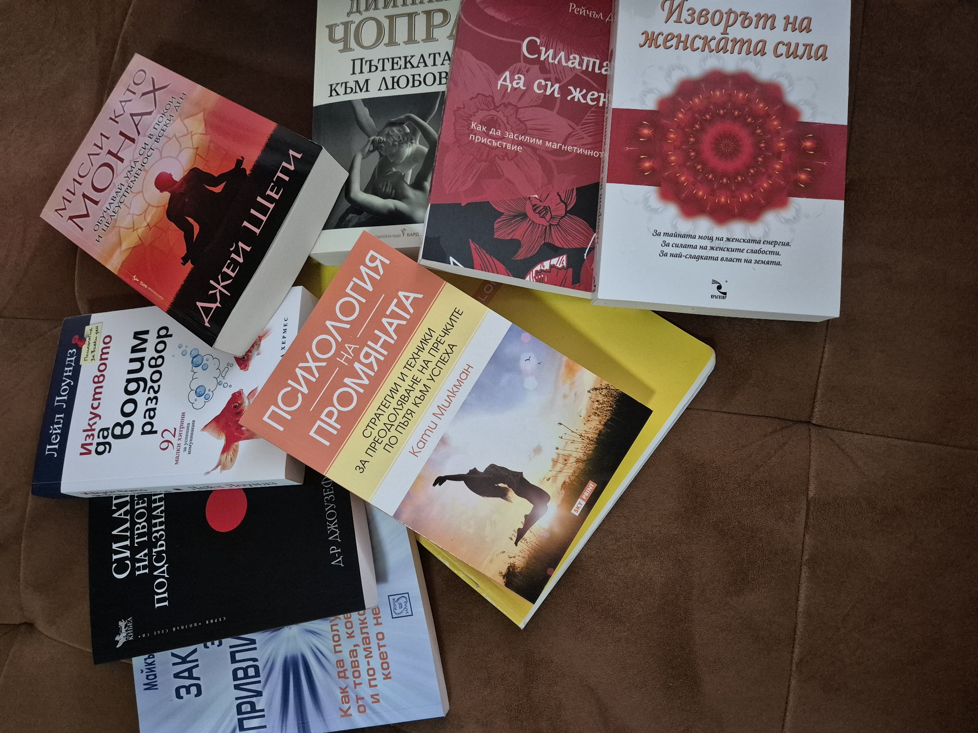 An overhead shot displays a collection of self-help and personal development books in Bulgarian, scattered across a textured, brown upholstered surface, possibly a sofa or armchair. The books are positioned at various angles, suggesting a casual arrangement.
Visible titles and authors include:
* "МИСЛИ КАТО МОНАХ" (Think Like a Monk) by ДЖЕЙ ШЕТИ (Jay Shetty), with the subtitle "ОБУЧАВАЙ УМА СИ В ПОКОЙ И ЦЕЛЕУСТРЕМЕНОСТ ВСЕКИ ДЕН" (Train your mind in peace and purpose every day).
* A book by ЧОПР (Chopra), titled "ПЪТЕКАТА КЪМ ЛЮБОВ" (The Path to Love).
* "Силата Да си жена" (The Power of Being a Woman) by Рейчъл Д (Rachel D.), subtitled "Как да засилим магнетичното присъствие" (How to enhance magnetic presence).
* "Изворът на женската сила" (The Source of Female Power), which mentions "За тайната мощ на женската енергия. За силата на женските слабости. За най-сладката власт на земята." (For the secret power of female energy. For the power of female weaknesses. For the sweetest power on Earth).
* "Изкуството да Водим разговор" (The Art of Conversation) by Лейл Лоундз (Leil Lowndes), offering "92 малки хитрини за успешна комуникация" (92 small tricks for successful communication).
* "ПСИХОЛОГИЯ НА ПРОМЯНАТА" (Psychology of Change) by Кати Милкман (Katy Milkman), covering "СТРАТЕГИИ И ТЕХНИКИ ЗА ПРЕОДОЛЯВАНЕ НА ПЪТЯ КЪМ УСПЕХА" (Strategies and techniques for overcoming the path to success).
* "СИЛАТА НА ТВОЕТО ПОДСЪЗНАНИЕ" (The Power of Your Subconscious Mind) by Д-Р ДЖОУЗЕФ (Dr. Joseph).
* A partially visible book titled "ЗАКОНЪТ НА ПРИВЛИЧАНЕТО" (The Law of Attraction) by Майкъ (Michael), with text "Как да получите повече от това, което искате и по-малко от това, което не искате" (How to get more of what you want and less of what you don't want).
The overall theme of the books revolves around personal growth, self-empowerment, and communication. The lighting is bright and even, indicating an indoor setting.