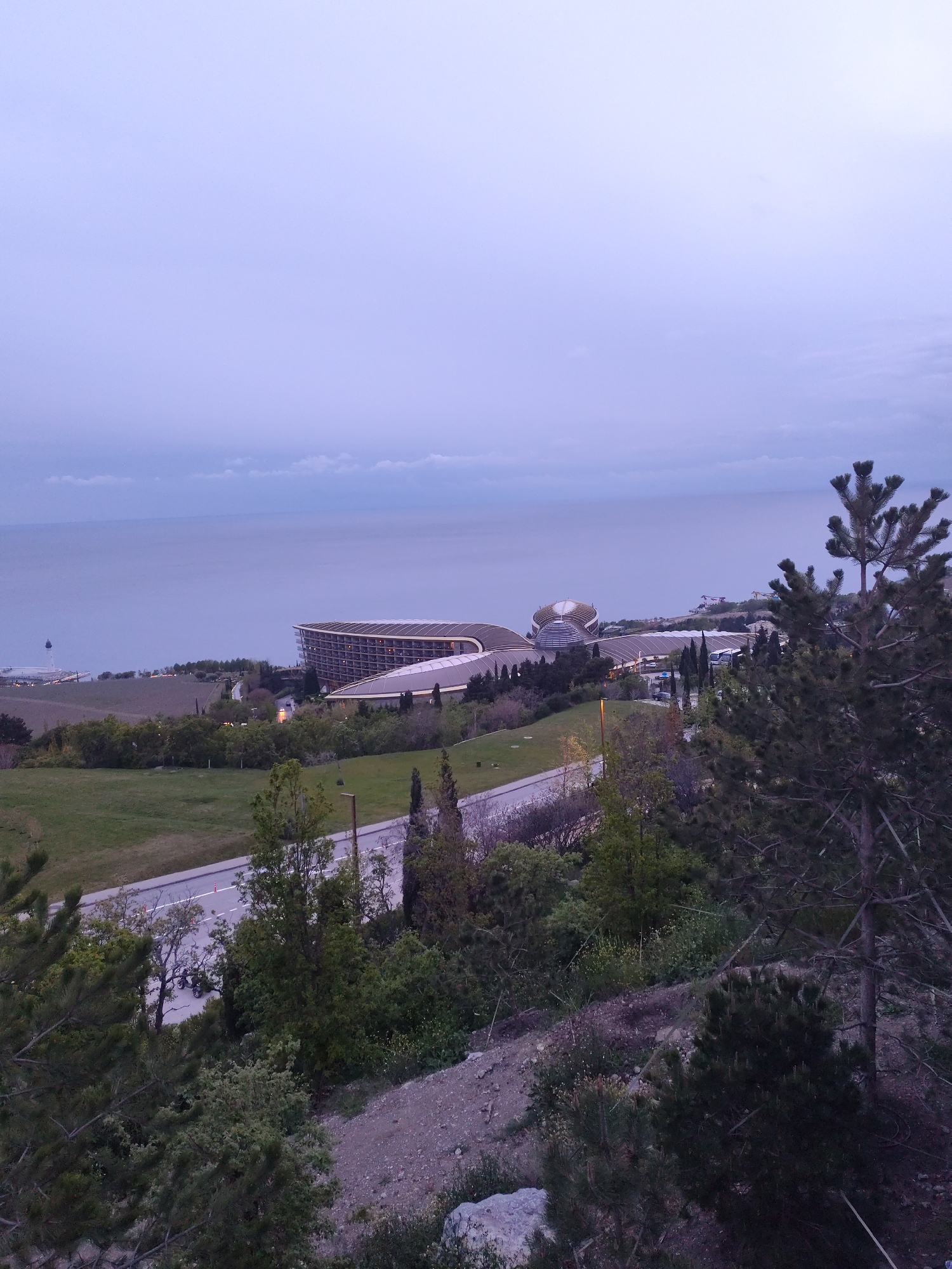 The image captures a panoramic view of a coastal landscape, likely within the area of Симеизский поселковый совет, Ukraine. The scene is observed from an elevated perspective, looking down towards the sea and a developed coastline at twilight or dawn.
The upper portion of the frame is dominated by a vast, soft purple-blue sky with visible cloud cover, indicating either the early morning or late evening hours. Below the sky, the calm, grey-blue sea stretches across the middle distance, meeting the land.
Along the coastline, prominent modern architectural structures with distinctive curved roofs and multiple illuminated windows suggest a resort or hotel complex. To the far left on the distant coastline, a slender lighthouse or tower is visible. The mid-ground features a mix of green fields, darker patches of vegetation, and scattered trees. A road winds through this landscape, with a street light faintly visible near it.
The foreground is characterized by lush coniferous trees, with a large, dark pine tree framing the right side of the image. The immediate foreground also shows a rocky, uneven slope with sparse greenery. No specific activities or people are discernible. The overall atmosphere conveyed by the dim, diffused light is one of tranquility and serenity. No visible text is present in the image.