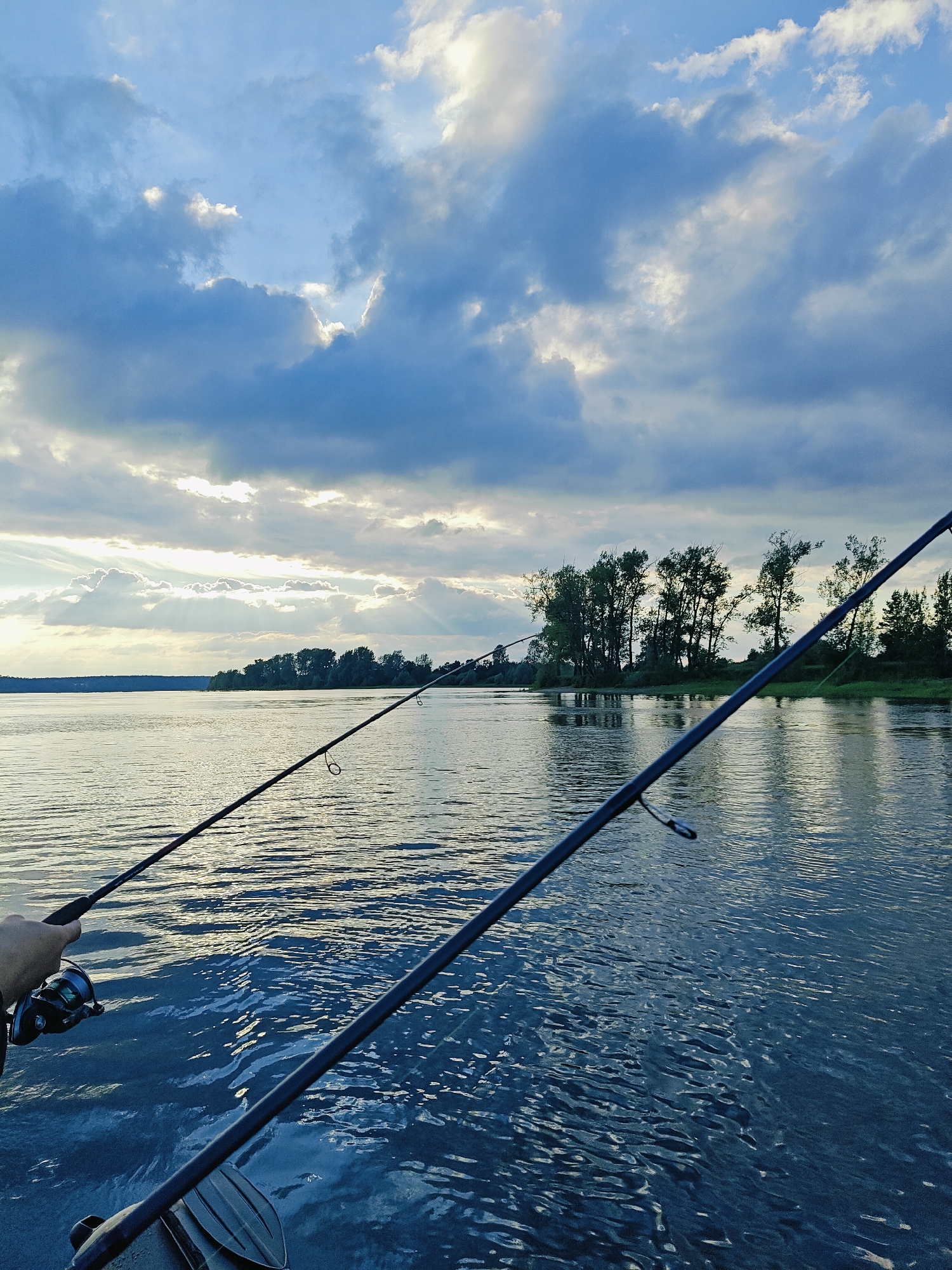 This media file depicts a scenic view of a river with two fishing rods extended into the water. A person's hand is visible holding one of the fishing rods, which has a reel attached. The scene is set during what appears to be either sunrise or sunset, given the dramatic lighting with sun rays piercing through the clouds. The sky is filled with a mix of light and dark clouds, creating a dynamic and atmospheric backdrop.
The river occupies the majority of the frame, its surface rippling with gentle waves that reflect the subdued light. In the distance, a lush, tree-lined shore creates a natural border to the river. The trees are dense, suggesting a natural or rural environment.
The overall mood is peaceful and serene, evoking a sense of calm and connection with nature. The activity clearly taking place is fishing, a leisurely pursuit often enjoyed in such natural settings. The location context provided, Ol'khovskoye, Russia, further reinforces the impression of a tranquil, possibly remote, natural landscape. There is no visible text within the image.