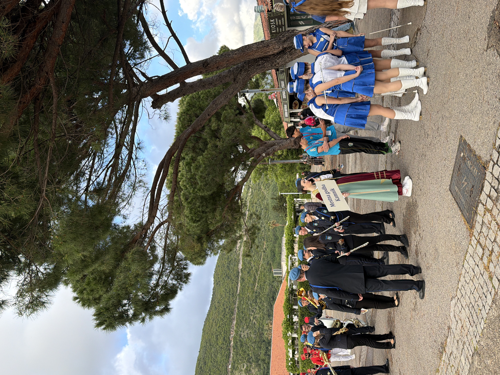 The image captures a musical parade in the city of Budva, Montenegro. The parade consists of two main groups: a marching band and a baton twirling team. The marching band members are dressed in dark uniforms with blue hats and are carrying various musical instruments, including clarinets, trumpets, and saxophones. The baton twirling team is dressed in blue and white uniforms with white boots. A person in traditional Montenegrin clothing stands next to the parade with a sign that reads "Mestna godba Kamnik". The parade is taking place on a paved road adjacent to a building with a red tile roof. In the background, there is a green, mountainous landscape and a partially visible sky. There is a large tree with dense foliage casting a shadow on the scene. The sign "KIOSK" is visible in the background.