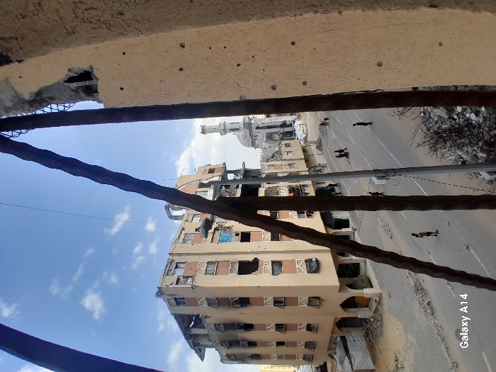 The image depicts a street scene in Khan Yunis, Palestinian Territory, characterized by extensive structural damage to urban buildings, viewed from an elevated position, possibly from within a damaged structure.
In the immediate foreground, two thick, rusted rebar elements diagonally cross the frame, partially obstructing the view. To the far right, a section of a damaged concrete wall exhibits multiple small impact marks, consistent with bullet holes or shrapnel. Below this, a pile of rubble and twisted metal debris is visible on the ground. A utility pole with wires stands upright to the right of the left rebar.
The midground features a two-lane asphalt road running from the foreground into the distance. On the left side of the street, a multi-story building, approximately five to six stories tall, shows severe damage. Numerous windows are blown out, revealing dark interior spaces. The facade exhibits multiple impact holes, shattered sections, and evidence of charring, particularly on the lower floors. Parts of the building's structural elements appear exposed.
On the street itself, several individuals are present. Approximately seven to ten people are visible, mostly walking. One person in a yellow shirt stands alone in the middle of the left lane of the road. Further down the street, near the right sidewalk, a small group of three to four people are walking. Other individuals are distributed along the road.
In the background, at the end of the visible street, stands a mosque with a prominent, slender white minaret, appearing partially damaged. The surrounding buildings in the background also show signs of destruction. The sky overhead is a clear blue with scattered white clouds, indicating daylight conditions. The entire scene conveys a widespread state of urban devastation.