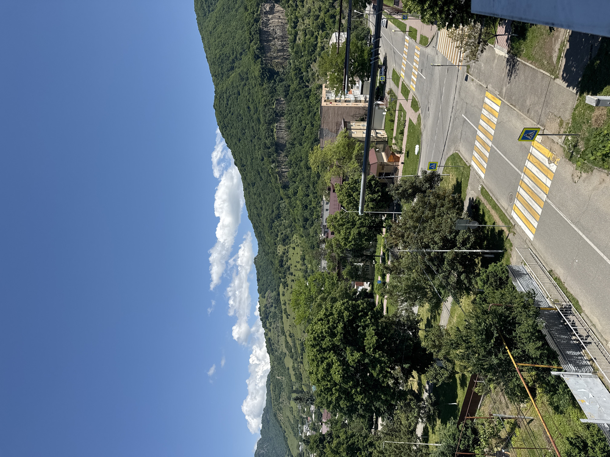 The image depicts an urban landscape with a clear blue sky and scattered white clouds. In the foreground, a paved road features multiple marked pedestrian crossings. On the sides of the road, there are sidewalks, grassy areas, and various trees. Buildings, including apartment blocks and smaller structures, are visible in the middle ground. Further in the distance, rolling green hills dominate the background, with some areas showing rocky outcrops. The overall scene suggests a sunny day in a city located in a hilly or mountainous region. The presence of pedestrian crossings, roads, and buildings indicates a developed area.