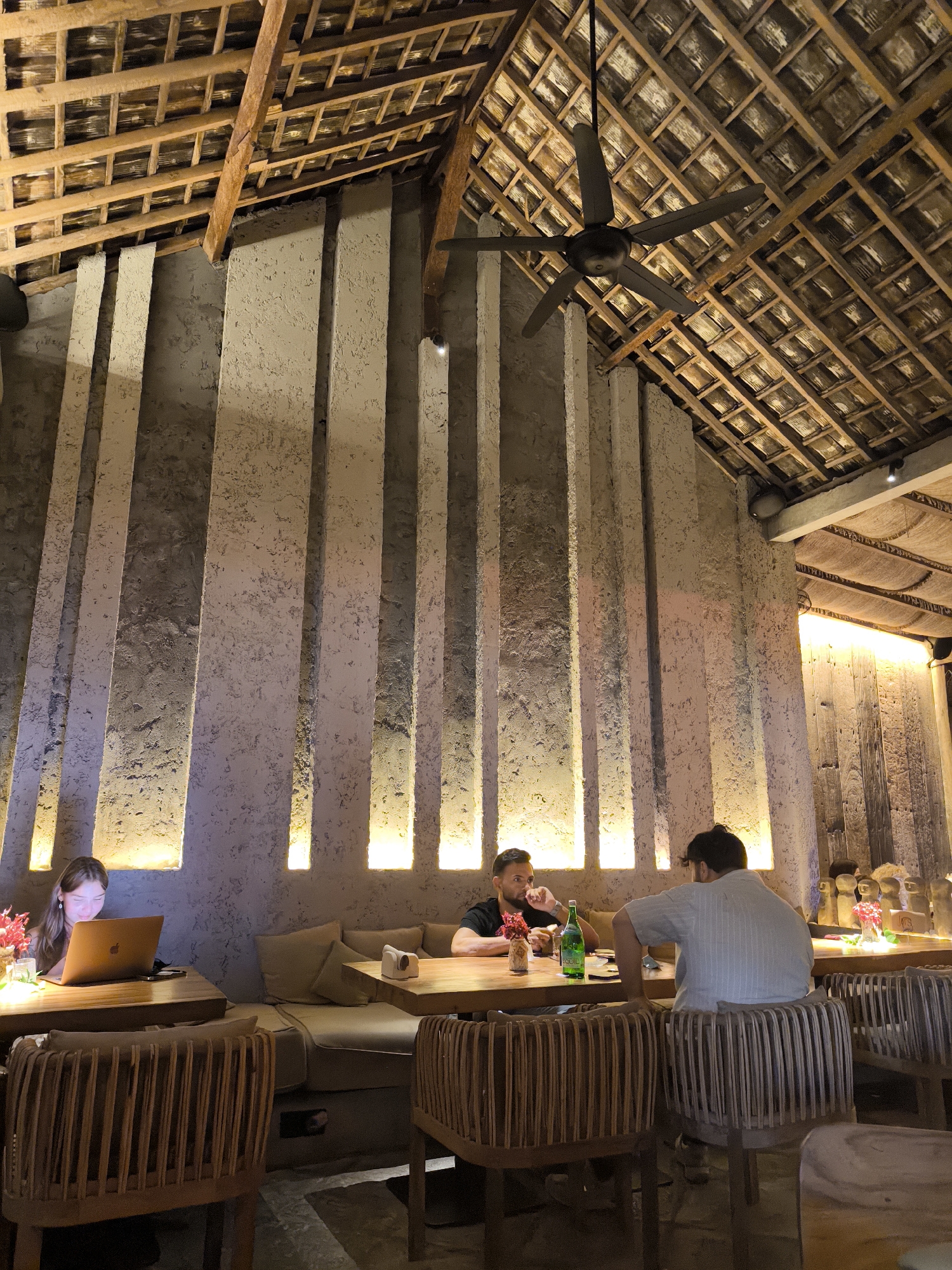This image captures the interior of a dimly lit restaurant or cafe, presenting a warm and inviting atmosphere. The dominant architectural feature is a high, vaulted ceiling constructed from exposed wooden beams and what appears to be woven thatch or bamboo material. A dark ceiling fan hangs centrally from this rustic ceiling.
The walls are distinctive, featuring tall, textured, light-colored vertical panels. Recessed lighting installed at the base of these panels casts a warm yellow glow upwards, highlighting the texture and creating striking vertical lines of illumination.
Three individuals are visible within the space. On the left, a woman with long hair is seated at a wooden table, intently focused on a glowing laptop screen. In the mid-ground, two men are seated at another wooden table. One man, wearing a black t-shirt, faces left, appearing to be engaged in conversation or contemplation, with a green bottle of water (labeled "EQUIL") and a small vase of red flowers on the table before him. The second man, in a light-colored shirt, has his back mostly to the camera.
The furniture consists of sturdy wooden tables and chairs with slatted, curved backs, complemented by bench seating with plush cushions along the wall. The overall ambiance is relaxed and comfortable, suggesting an evening setting given the reliance on artificial lighting. The architectural style, particularly the thatched roof, aligns with a tropical location like North Kuta, Indonesia.