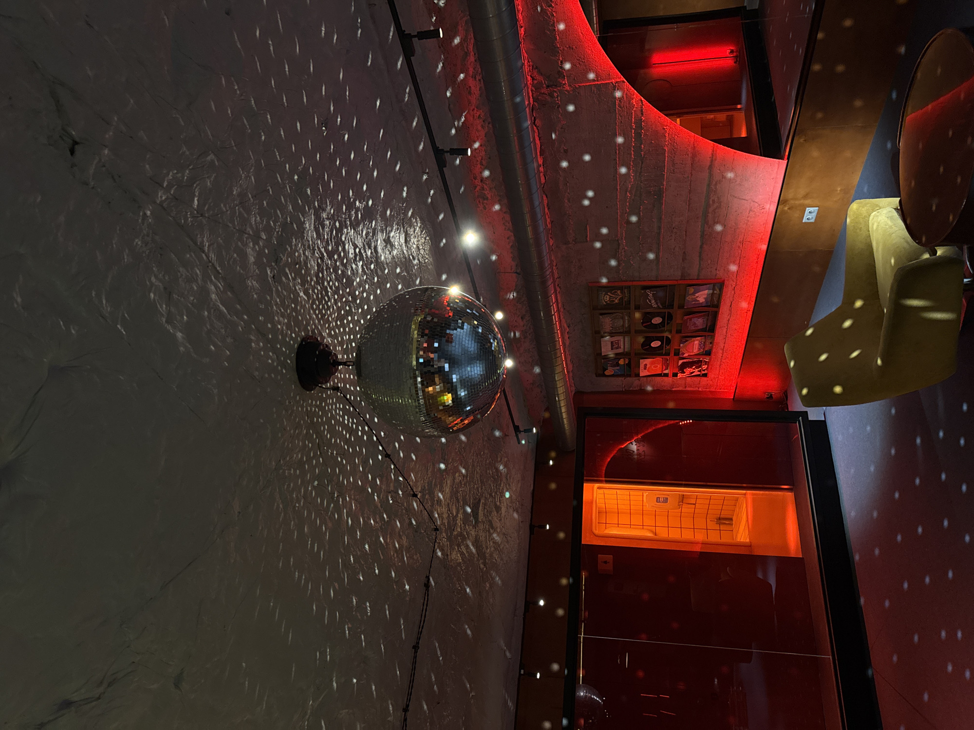 A disco ball hangs from a wall, scattering light patterns across the surrounding surfaces. The walls and ceiling appear to be textured, possibly concrete or plaster. A series of lights are mounted along the walls. The room is dimly lit with red lighting. In the background, a shelf of records and an open doorway are visible. A yellow chair sits on the right side of the frame, partially obscured. It is indoors. There is no indication of Yerevan, Armenia in the image.