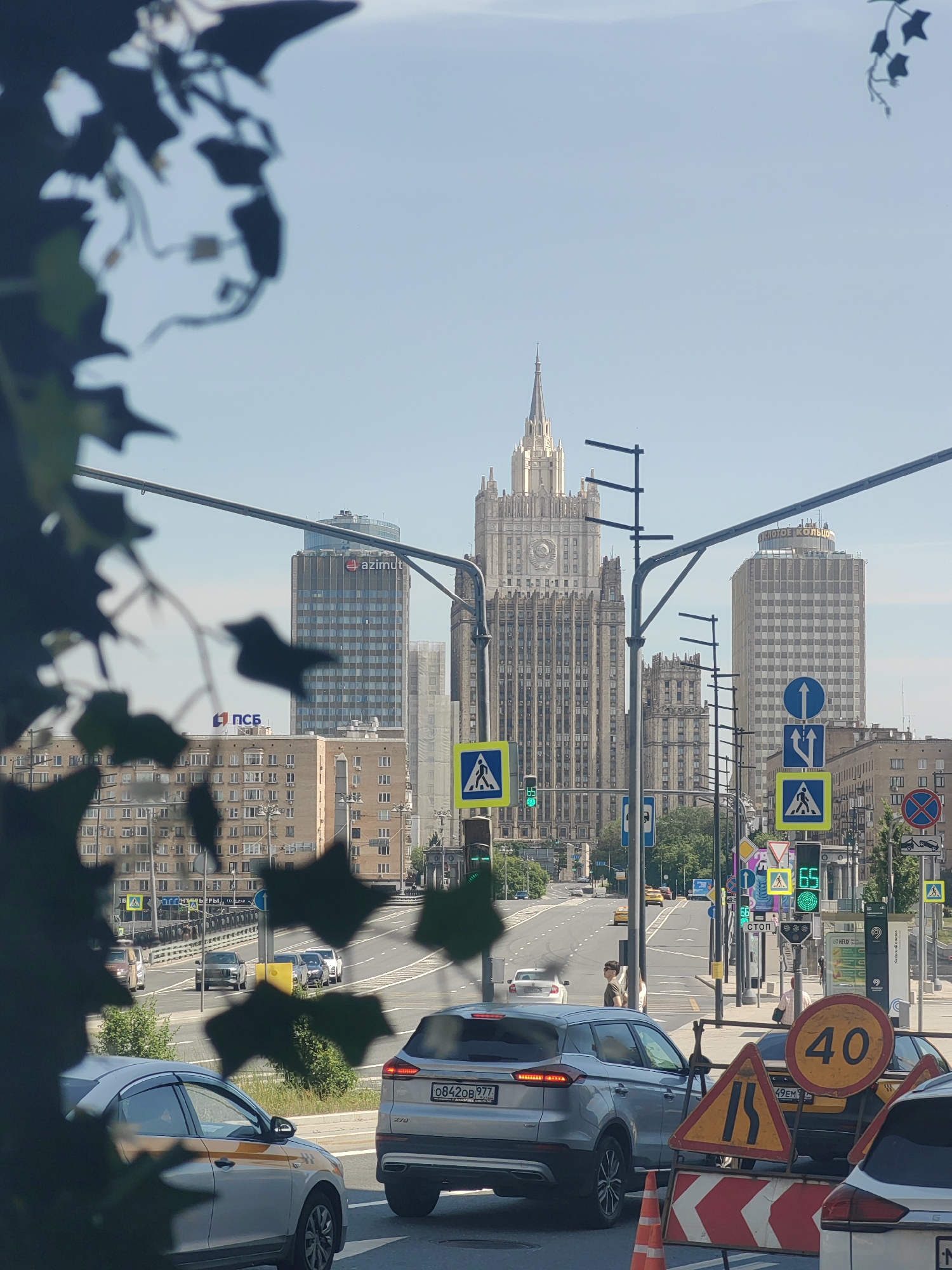 This image captures a daytime urban street scene in Moscow, Russia, looking towards Smolenskaya Square. The prominent Stalinist-era skyscraper in the background is the Main Building of the Ministry of Foreign Affairs of Russia.
In the immediate foreground, dark green foliage, possibly from a tree, is out of focus and frames the left and upper portions of the image. The scene centers on a multi-lane asphalt road. In the lower center, a silver Geely Atlas (or Proton X70) SUV, bearing Russian license plate O842OB 977, is stopped, facing away from the viewer. To its left, the rear of a silver sedan with an orange stripe, indicative of a taxi, is partially visible. A white car is partially visible on the far right.
A cluster of temporary road signs and barriers is visible in the lower right: a circular yellow speed limit sign with "40" in black, a triangular yellow warning sign depicting a road narrowing, and a red and white striped barrier with an orange traffic cone.
Further down the road, several blue and white regulatory and informational road signs are mounted on poles and an overhead gantry. Two square blue and yellow pedestrian crossing signs are visible, one displaying a green light for pedestrians. Traffic lights along the road are predominantly green. A male pedestrian, wearing sunglasses and a dark t-shirt, stands on the right sidewalk near a crosswalk.
In the mid-ground, various buildings line the street. On the left, a multi-story building displays a red "ПСБ" (PSB) logo on its roof. Behind it, a modern, dark-colored high-rise building with a rounded top features a prominent "azimut" logo.
The background is dominated by the majestic Main Building of the Ministry of Foreign Affairs, a Stalinist skyscraper with a central spire, intricate facade details, and a Soviet emblem above its lower entrance. To the right of the MFA building, another tall, lighter-colored building is visible, with faint Cyrillic text, possibly "ОРЛОВОЕ КОЛЬЦО" or similar, near its roofline. A bridge or overpass with vehicles is visible in the mid-left background. The sky is clear and pale blue.
