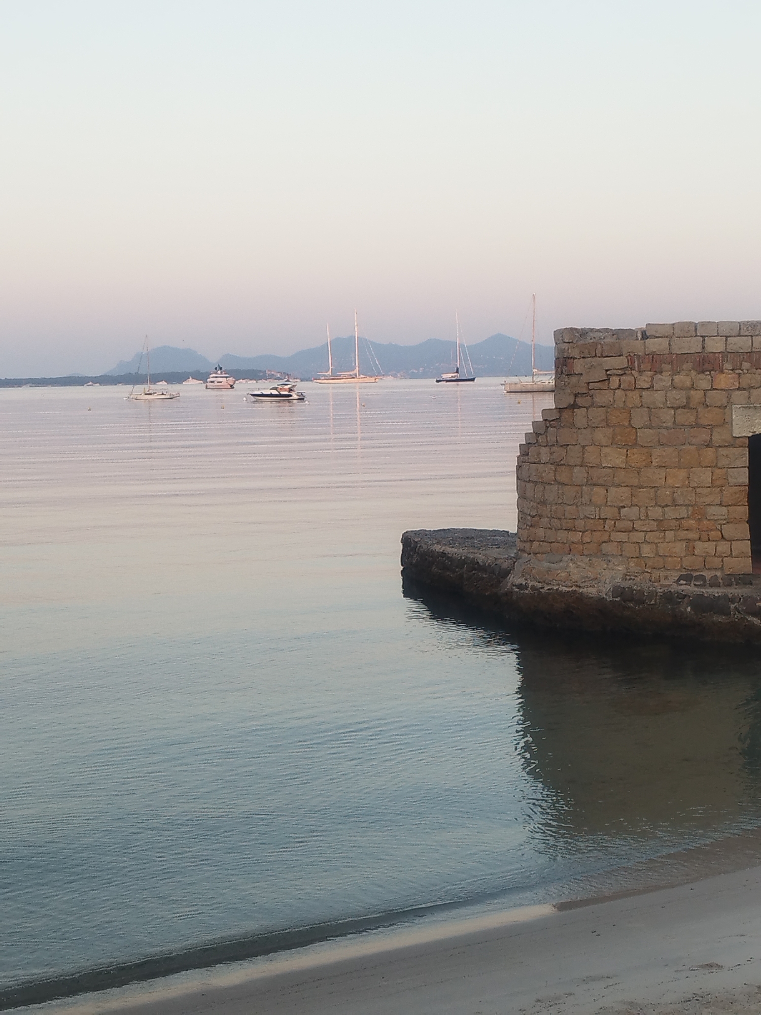 The image depicts a tranquil scene of a harbor in Antibes, France, likely at dawn or dusk given the soft, diffused light. In the foreground, a sandy beach transitions to calm, rippling water. To the right, a partially ruined stone structure, appearing to be an old sea wall or fortification, juts out into the water. Beyond this, several sailboats and yachts are anchored or moving slowly in the bay. In the distance, a hazy, mountainous coastline is visible under a pale sky. The overall atmosphere is serene and still, with reflections of the boats and the sky visible on the water's surface. There are no people visible in the image.