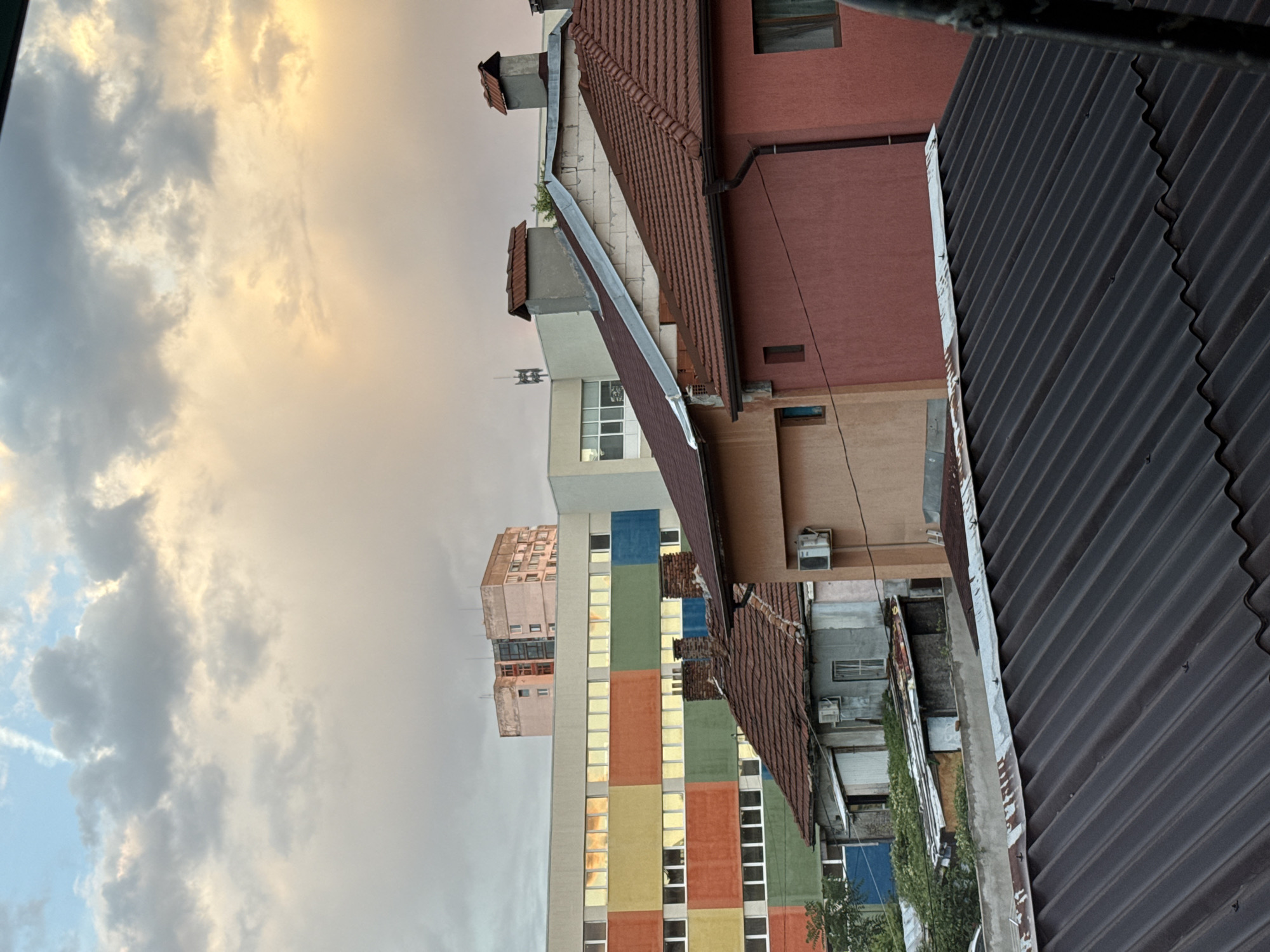 This is a photograph taken from a rooftop in Sofia, Bulgaria, looking towards a city skyline at dusk. The foreground is dominated by the corrugated metal roof of the viewpoint. Behind this, a row of houses with terracotta tiled roofs are visible, some showing signs of age and disrepair, with air conditioning units attached to their walls. Further back, a modern, colorful building with a facade divided into large squares of yellow, green, red, and blue glass is prominent. Above this, a taller, older, pale pink building with multiple windows stands. The sky is filled with dramatic, scattered clouds, illuminated by the warm hues of the setting sun, with hints of blue and white in the upper sections. The overall impression is one of urban density with a mix of old and new architecture under a dynamic sky.