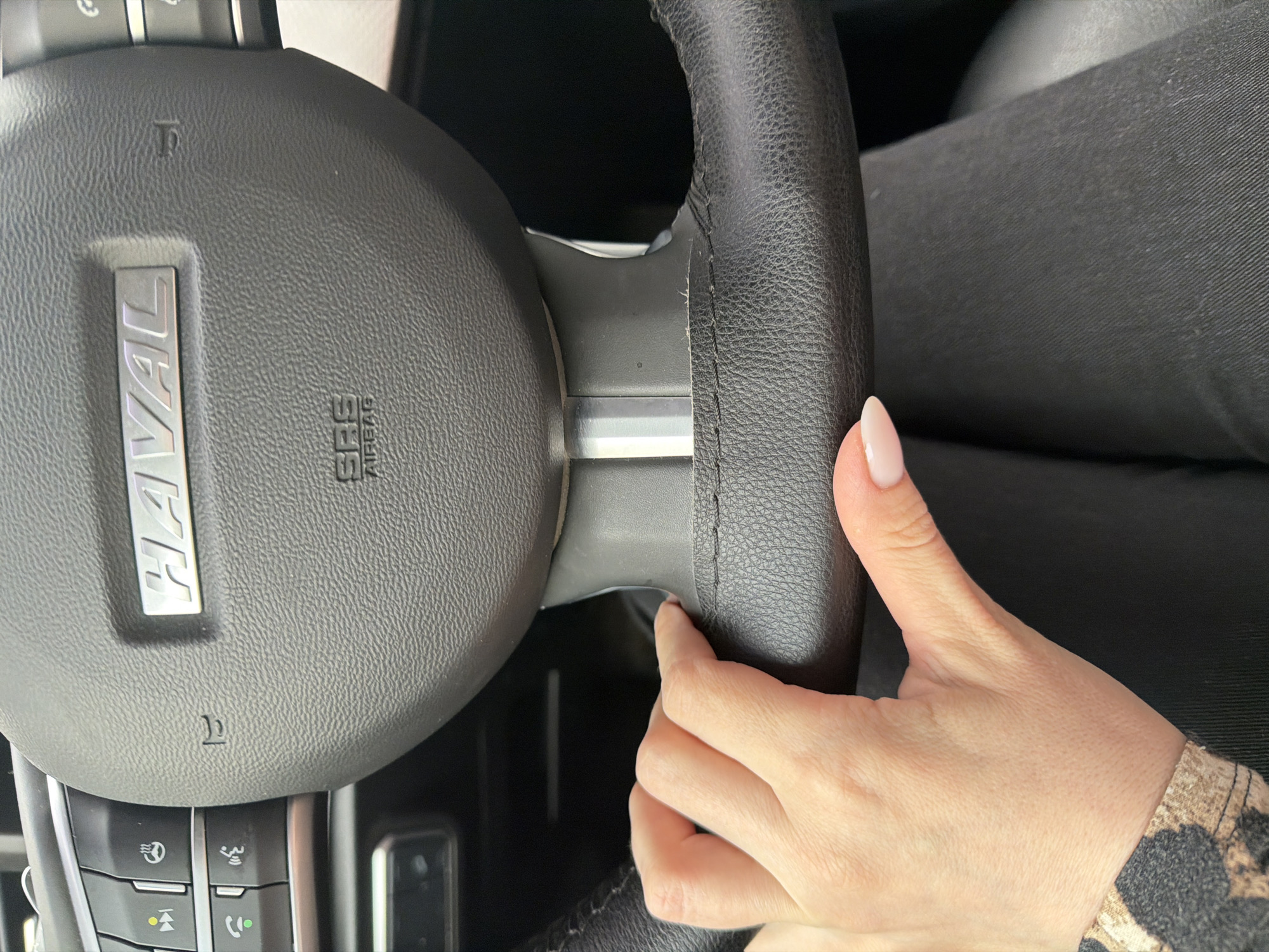 The image shows the steering wheel of a Haval car. The car's logo, "HAVAL," is visible on the central part of the steering wheel. The steering wheel is black, with silver accents. A person's hand with manicured fingernails rests on the wheel. The hand is fair-skinned and wears a printed sleeve. The background shows parts of the car's interior, including control buttons.
The location in the city of Izhevsk, Russia, cannot be determined from the image.