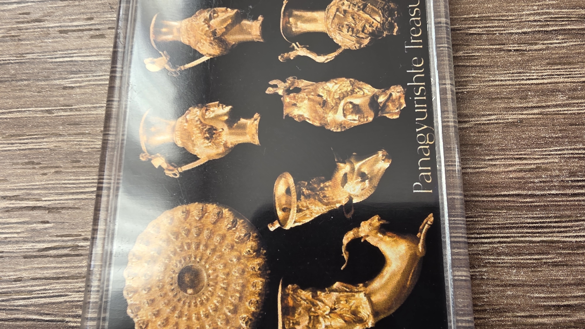 The image shows a pamphlet or card featuring a collection of golden artifacts on a black background, labeled "Panagyurishte Treasure." The artifacts are intricate and appear to be ancient gold vessels and decorative animal figures. The surface the card rests on is a brown, wood-grain pattern. The pamphlet may be from Blagoevgrad, Bulgaria, where the Panagyurishte Treasure has been displayed.