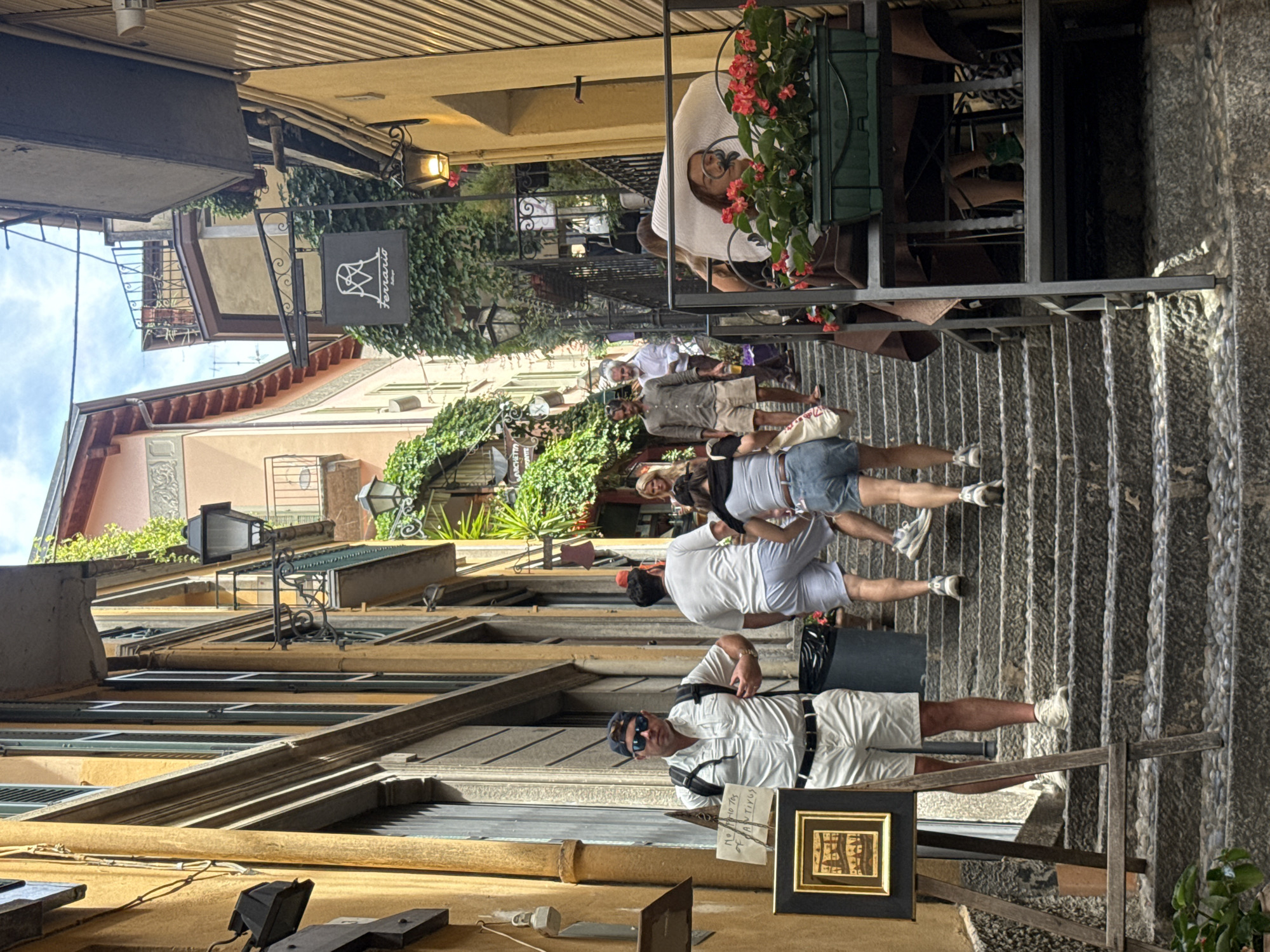 The image depicts a narrow, stone-paved staircase in a European city, possibly Italy. The buildings lining the staircase are tall and narrow, with yellow and pale pink facades adorned with green shutters and balconies. Lush green vines cascade down some of the buildings, creating a vibrant contrast with the architecture. Several people are visible on the stairs, some walking up and others standing.
In the foreground, a man wearing a white short-sleeved shirt, white shorts, sunglasses, and a baseball cap stands on the left side of the stairs, facing slightly away from the camera. He has a backpack on. Next to him, an easel holds a framed artwork with a dark frame and a small, detailed image inside. A sign with handwritten text is attached to the easel.
Further up the stairs, a group of people are ascending. One person in the center is wearing a white t-shirt and grey shorts, while another, to their left, wears a white t-shirt and light blue shorts. A woman with dark hair is walking up behind them, wearing denim shorts and a black top.
To the right of the staircase, a metal structure with a table and chairs is set up, possibly a seating area for a cafe or restaurant. A person wearing a large white sun hat is seated at the table, with their head tilted back. A planter filled with red flowers sits on the railing of this seating area.
A sign with the word "Ferrario" and a stylized logo hangs from the side of a building, adorned with more greenery. Street lamps are visible on some of the buildings. The overall atmosphere suggests a charming, sun-drenched European street scene.
