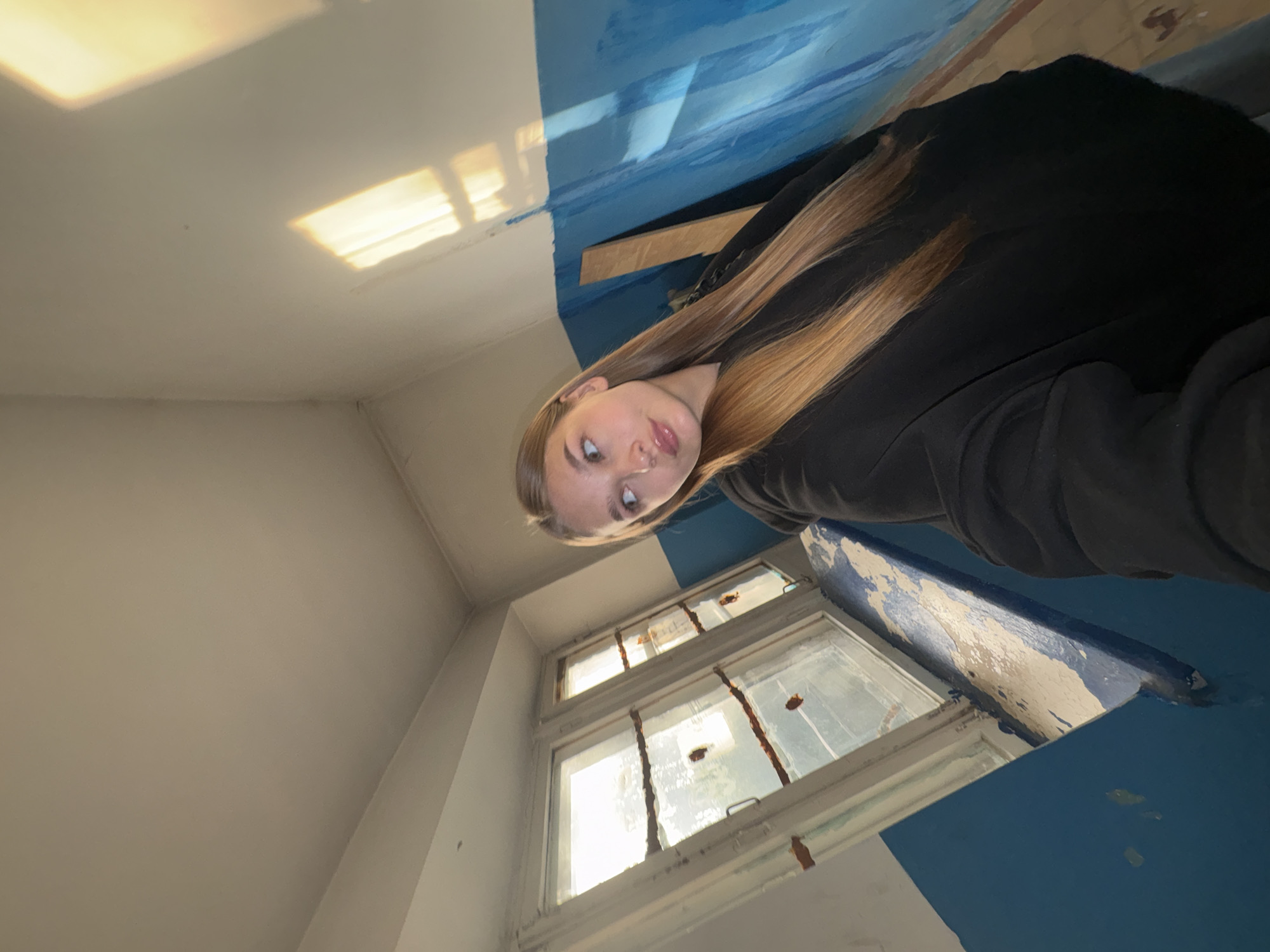 A young woman with long blonde hair is standing indoors, facing left, and looking away from the camera. She is wearing a black hoodie. The background consists of a white ceiling, a white-framed window with two panes, and a wall painted blue and white. Sunlight is streaming through the window, creating bright reflections on the ceiling and wall. There is a wooden plank leaning against the blue section of the wall. The overall environment appears to be an older building, possibly a residential apartment or communal space, located in the city of Chaikovsky, Russia.