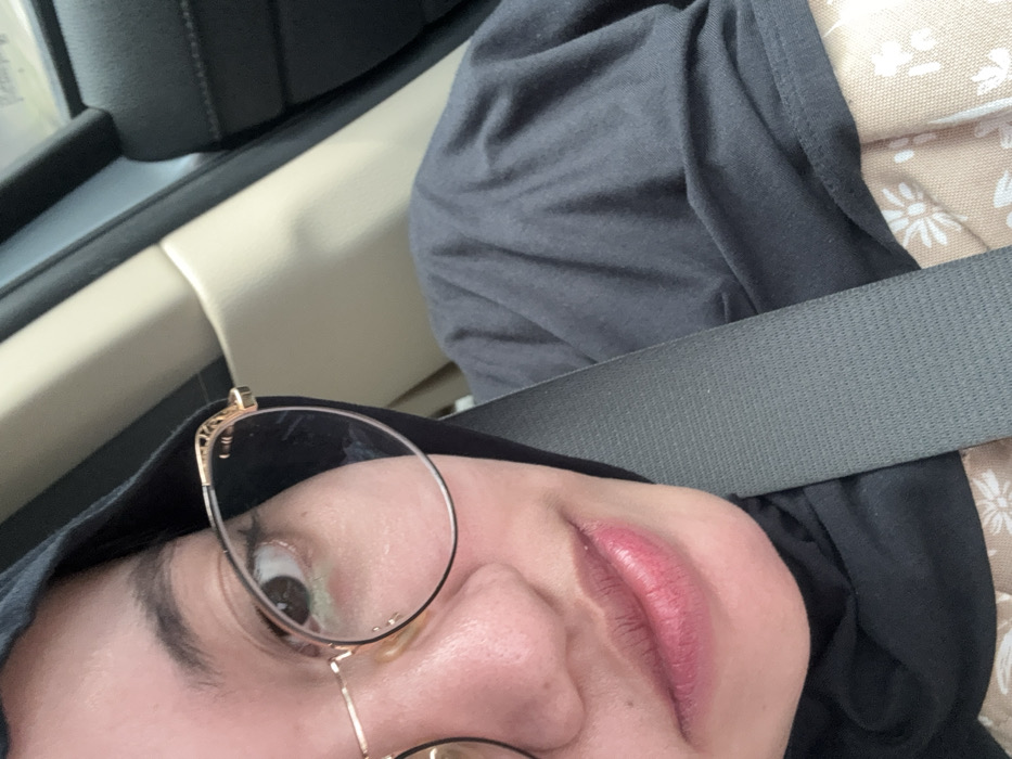 A close-up, angled selfie of a person wearing glasses and a dark head covering, possibly a hijab. The person is wearing a seatbelt. The background includes a car interior with a beige headrest and a black dashboard. The person's face is tilted towards the camera, showing one eye and part of the lips, which are pink. The glasses have thin, gold frames. The seatbelt is gray and positioned across the person's chest.