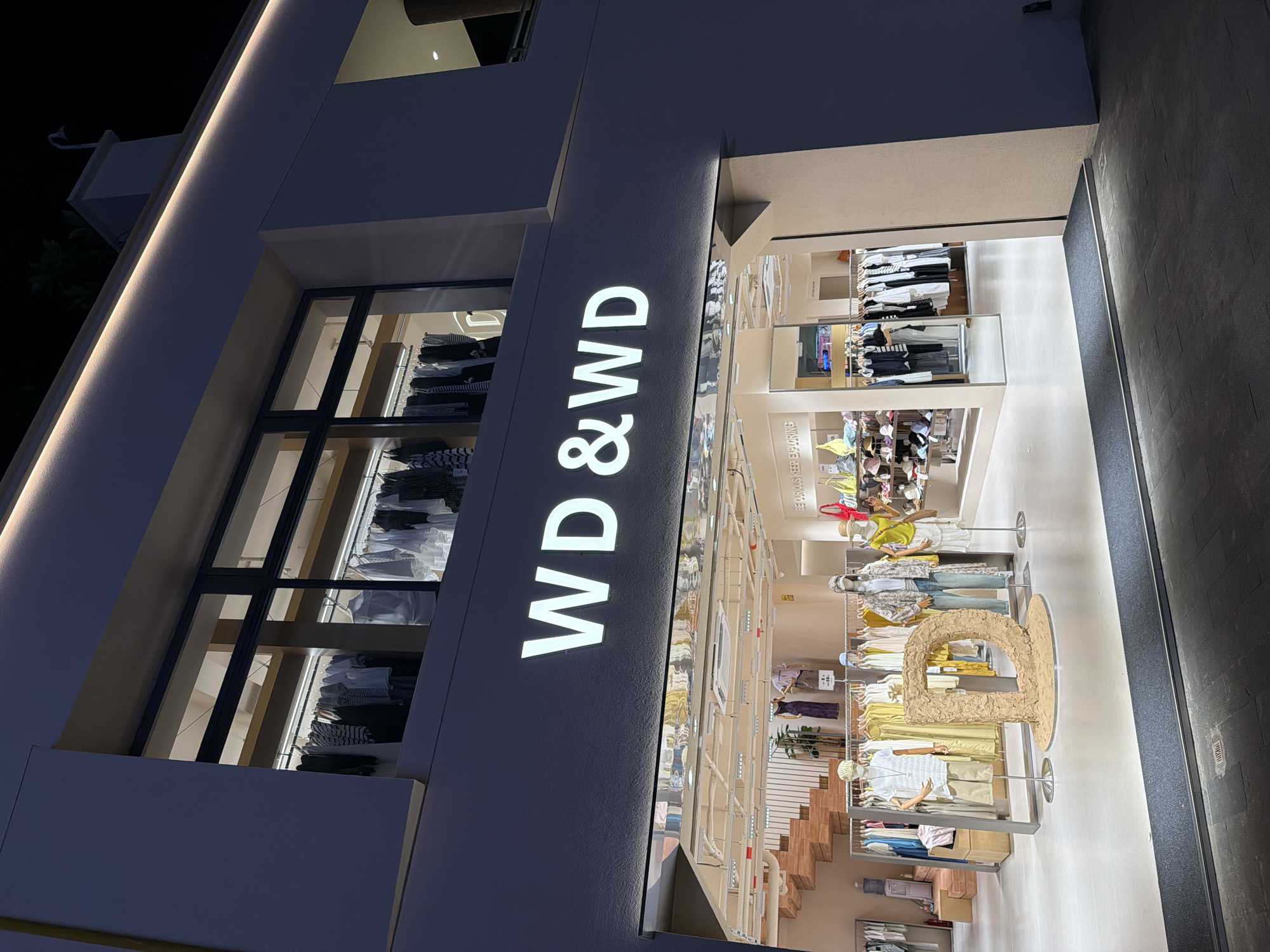 The image shows a daytime view of the "WD & WD" store, located in the Chongqing High-tech Zone, China. The store's name is prominently displayed on the facade in large, illuminated white letters. Through the large window, various clothing items are visible on racks and mannequins. The interior has a brightly lit, modern aesthetic. The front of the store opens directly onto a sidewalk.