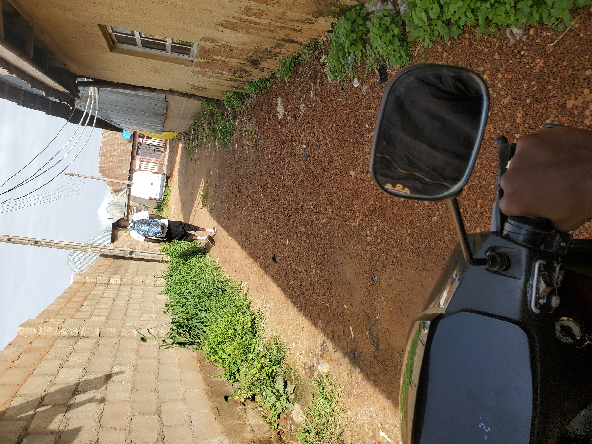 A person wearing a white t-shirt, black shorts, white socks, and dark sneakers walks away from the camera down a dirt path in Bukuru, Nigeria. The person carries a blue and black backpack. The path is flanked by a tall cinder block wall on the left and a yellow-painted building on the right. Green vegetation grows along the base of the wall and near the building. The path is covered in small brown pebbles and dirt. In the foreground, the handlebar and rearview mirror of a black motorcycle are visible, with a hand gripping the right handlebar. A shadow of a person and a motorcycle handlebar is cast across the path from the right. In the distance, a white van and other buildings with corrugated metal roofs are visible. Overhead, utility wires stretch across the sky.