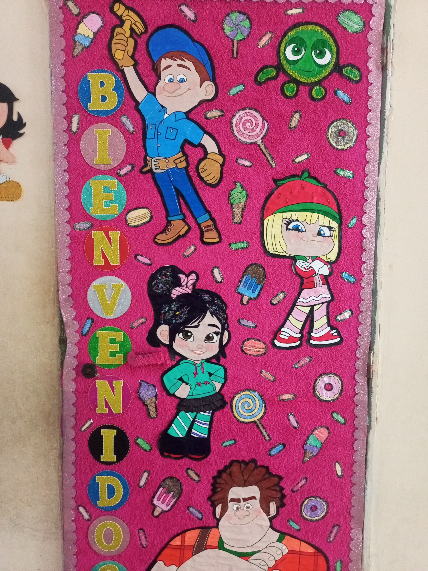 The image shows a colorful, handcrafted felt banner with characters from the movie Wreck-It Ralph. The banner is pink and decorated with various candy and dessert motifs like ice cream cones, donuts, and lollipops. It spells out "BIENVENIDO" vertically in large, circular letters in shades of blue, yellow, green, and red. The characters featured include Fix-It Felix Jr. at the top, Vanellope von Schweetz in the middle, and Wreck-It Ralph at the bottom. The banner is bordered with a sparkly pink scalloped trim and appears to be hanging on a wall. The environment suggests it is an indoor decoration, possibly for a party or a child's room. There is no visible indication of the city of Santa Ana, Venezuela in the image.