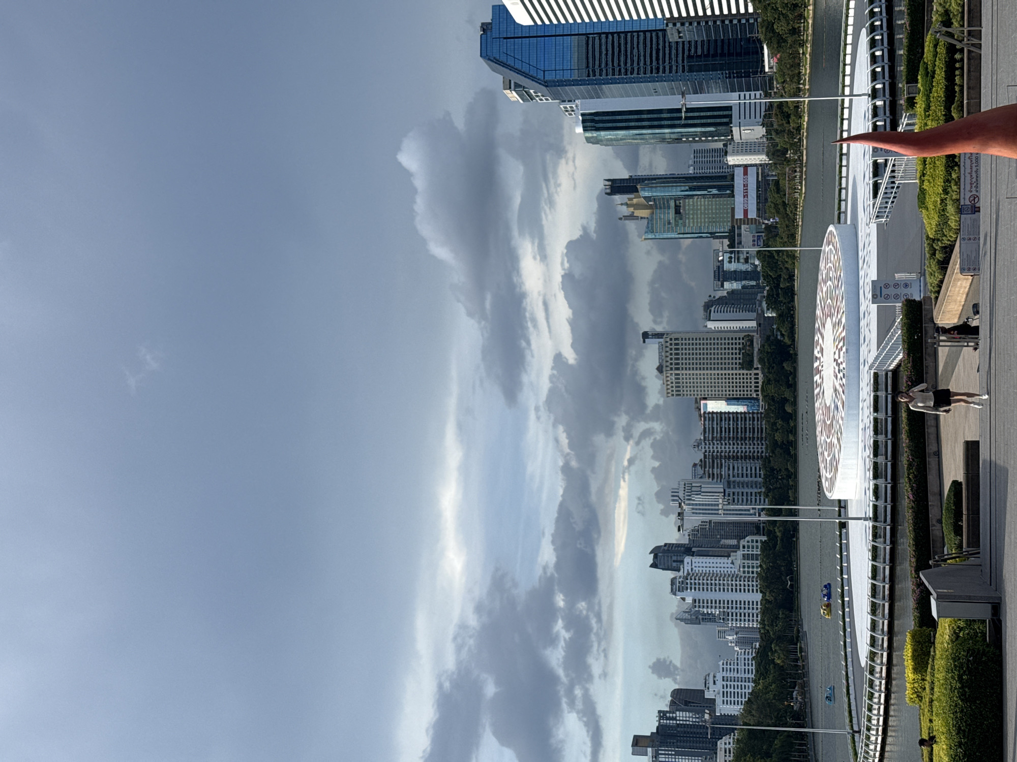 This media file captures a city skyline with a prominent body of water in the foreground, reflecting the overcast sky. The scene is dominated by a dense collection of modern skyscrapers, many of which have reflective glass facades, interspersed with lush green trees. A white, circular structure with intricate patterns is visible along the waterfront, suggesting a public space or attraction.
A person is seen on a pedestrian walkway bordering the water, appearing to be stretching or exercising. Further out on the water, small pedal boats are in motion. The sky is filled with dramatic, heavy clouds, with patches of brighter light indicating potential breaks in the overcast conditions. The overall lighting suggests it is daytime, possibly late afternoon or early evening, given the diffused light.
Visible text includes a partial advertisement banner on one of the buildings with the phone number "0899-111-555". Another sign with Thai script is present near the walkway. The presence of modern high-rise buildings and the distinct architectural style of the waterfront structure, along with the visible text in Thai, strongly suggest the location is Bangkok, Thailand. The atmosphere feels somewhat dramatic due to the cloudy sky, but the presence of people engaged in leisurely activities on the waterfront adds a sense of calm and urban life.
