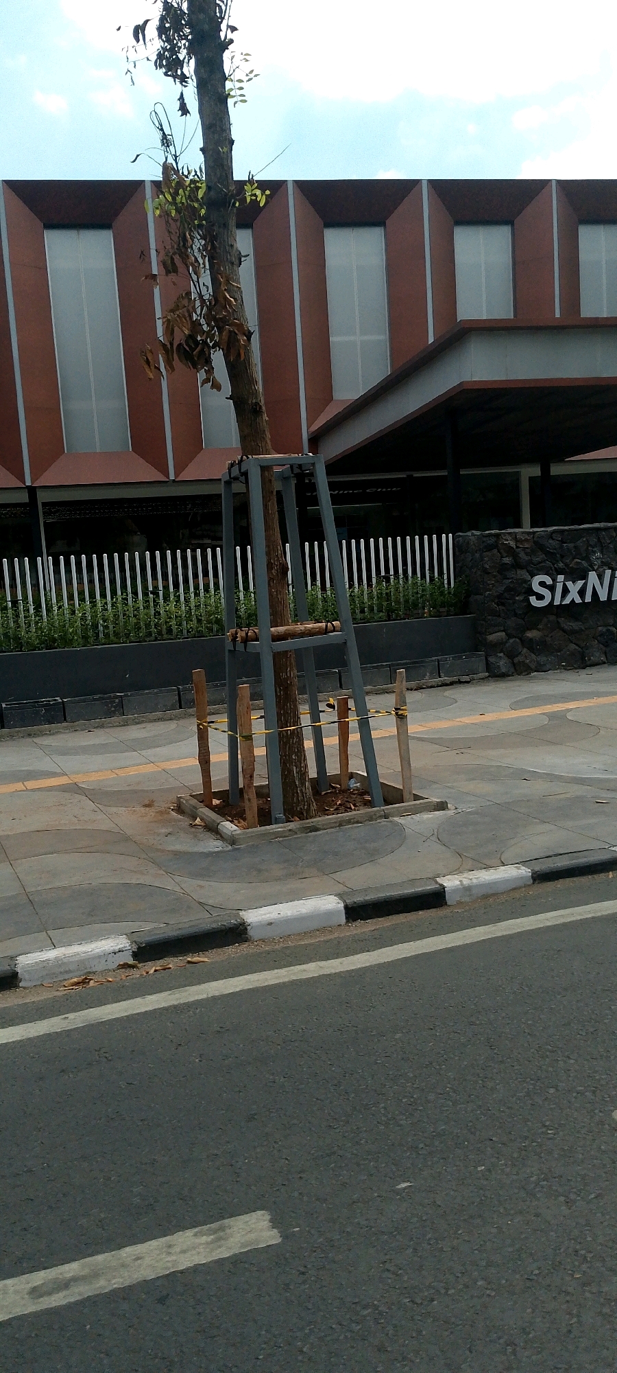 This is a medium shot, eye-level photograph taken from a street, looking towards a building and sidewalk. The foreground features a section of asphalt road with white lane markings, a raised concrete curb with alternating black and white paint, and a concrete sidewalk with a wavy pattern. A young tree, supported by a metal frame and wooden stakes, is planted in a square enclosure on the sidewalk. Yellow caution tape is visible tied around the wooden stakes supporting the tree. In the mid-ground, behind the sidewalk, is a row of white metal fencing, followed by a strip of green vegetation. Beyond this, a multi-story building with a distinctive facade of alternating brown and frosted glass sections is visible. A covered walkway extends from the building, and a stone wall with the white text "SixNi" is located to the right of the building's entrance. The sky above is partially cloudy. The scene is outdoors in an urban environment, likely a commercial area, given the building's architecture and signage. The location is identified as Bandung City, Indonesia.