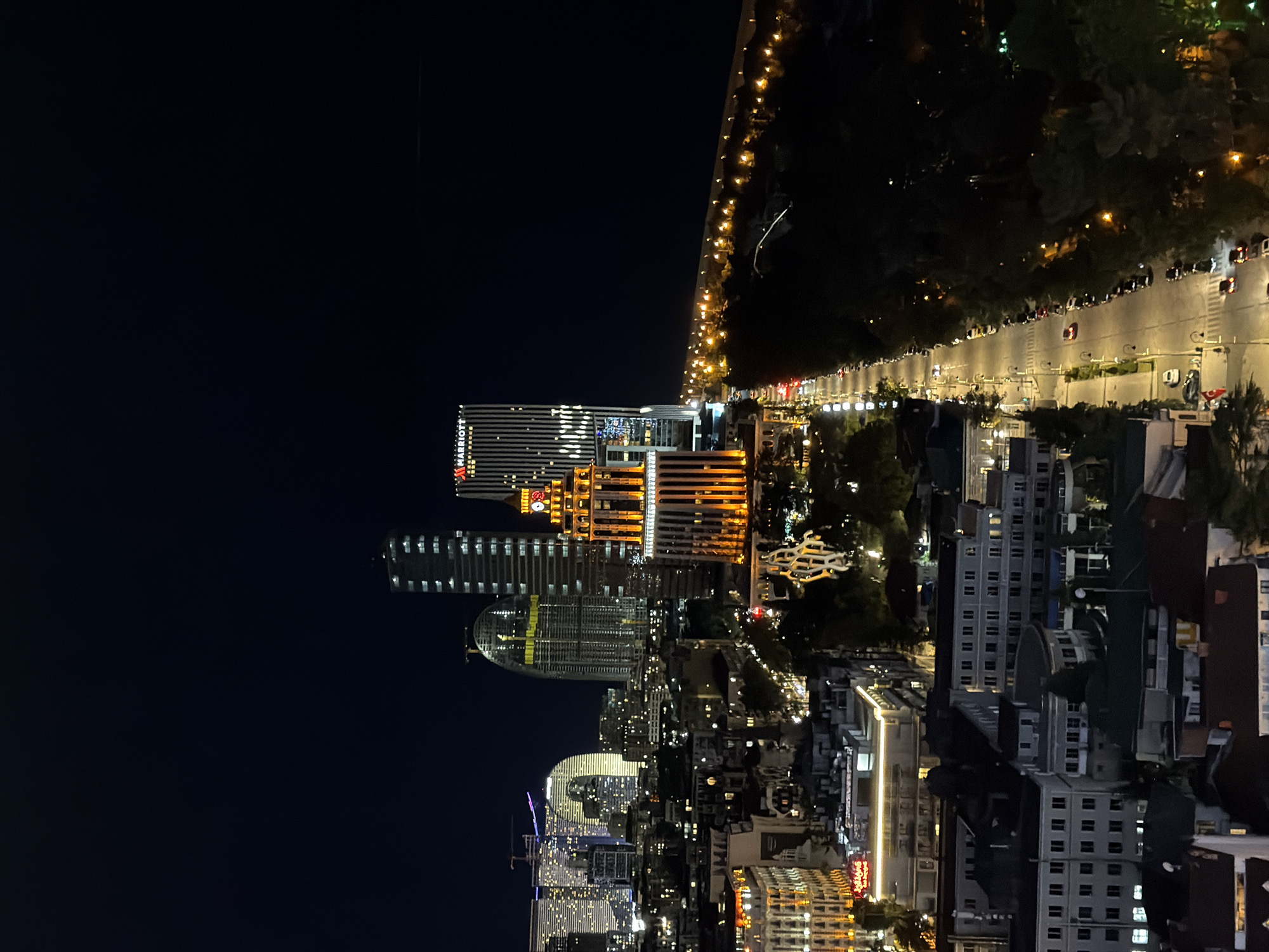 The image shows an aerial view of a city at night. Prominent landmarks include the Batumi Marriott Hotel, identifiable by its "Marriott" signage and distinctive architecture, and other modern high-rise buildings, some with visible construction, possibly including the Batumi Riviera Hotel. The environment includes illuminated streets with visible traffic, indicating urban activity. The overall setting suggests the image was taken in Batumi, Georgia, likely near the coastal area with modern development.